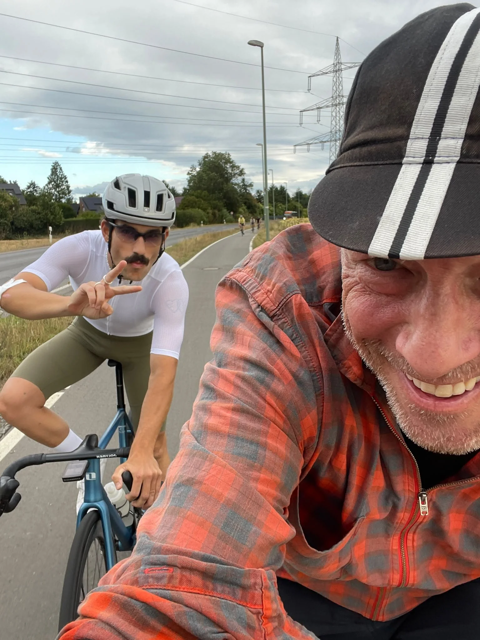Zwei Männer auf einem Radfahrer unterwegs auf der Straße, einer trägt einen Fahrradhelm und Sonnenbrille, der andere trägt eine Mütze und ein kariertes Hemd, im Hintergrund sind Bäume, Häuser und ein Himmel mit Wolken zu sehen.