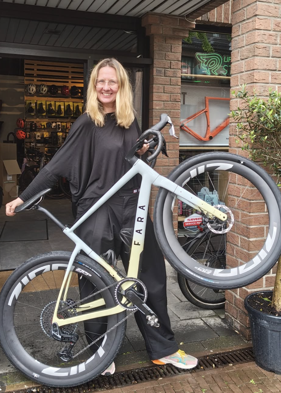 Eine Frau steht vor einem Fahrradgeschäft und hält ein modernes Fahrrad mit einem großen Reifen in der Hand, das sie aufrecht hält. Sie lächelt in die Kamera. Das Fahrrad ist schwarz und gelb, mit der Aufschrift ‚FARA‘. Hinter ihr sind Fahrradhelme, Fahrradteile und ein Rahmen eines Fahrrads an der Wand zu sehen, sowie ein Neonzeichen im Fenster.