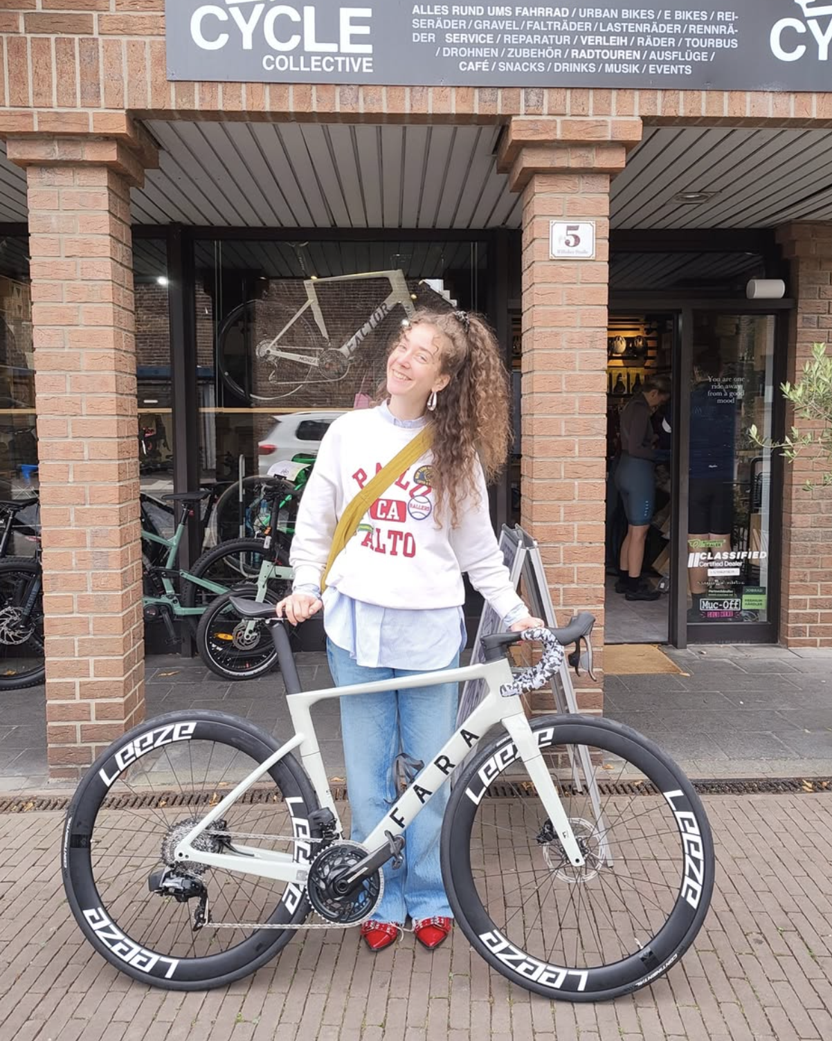 Eine junge Frau mit lockigem Haar, die auf einer Straße vor einem Fahrradgeschäft steht, neben einem weißen Fahrrad mit schwarzen Rädern, die Marke Leerze. Sie trägt eine weiße Sweatshirt mit „PALO ALTO“ bedruckt, blaue Jeans, rote Schuhe und einen gelben Schultergurt. Im Hintergrund sind Fahrräder im Geschäftsglasdisplay sichtbar. Die Fassade des Geschäfts ist aus roten Ziegeln gebaut, mit einem hohen Fenster und einer Tür.
