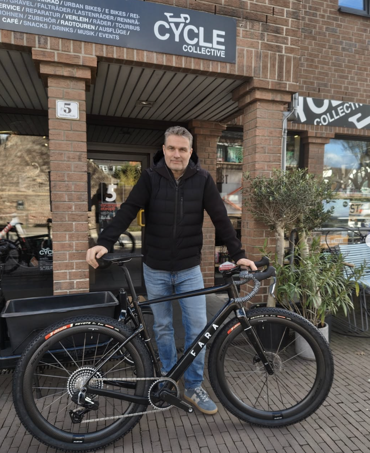 Ein Mann steht draußen vor einem Fahrradladen namens 'Cycle Collective' mit einem schwarzen E-Bike. Er trägt eine schwarze Jacke und blaue Jeans.