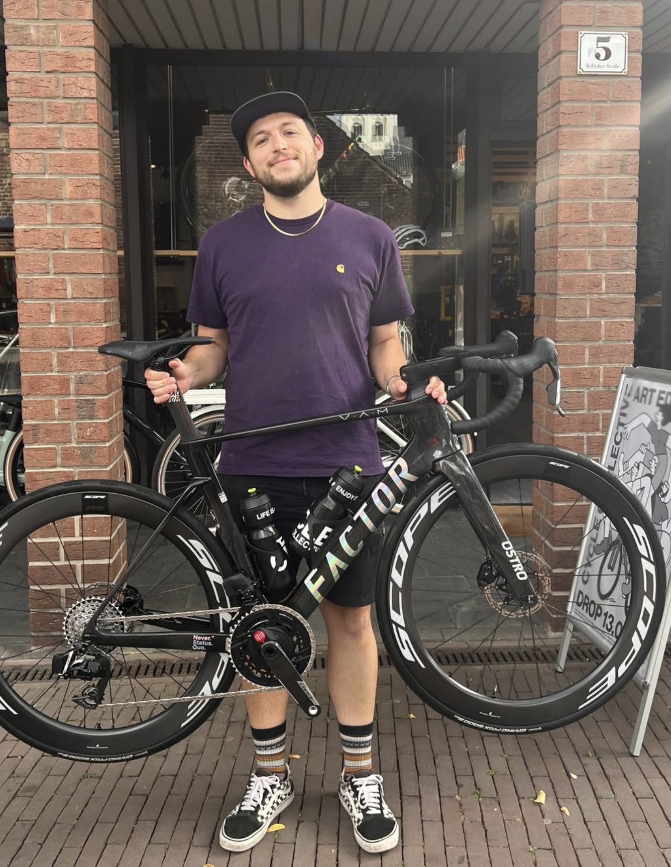 Junger Mann steht vor einem Fahrradgeschäft mit seinem Rennrad, trägt ein lila T-Shirt, Shorts, Sneakers und hält das Fahrrad am Lenker. Er hat einen Bart, trägt eine Kappe und eine Kette.