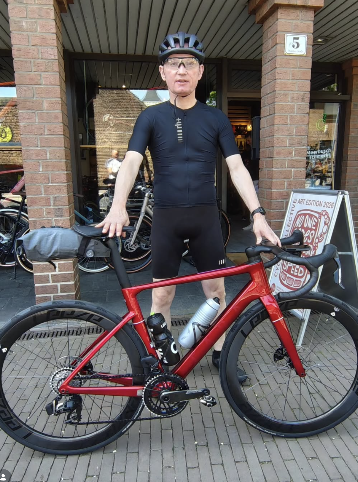 Ein Mann in Fahrradkleidung mit Helm steht vor einem roten Rennrad vor einem Geschäft. Im Hintergrund sind andere Fahrräder und eine große Werbetafel.