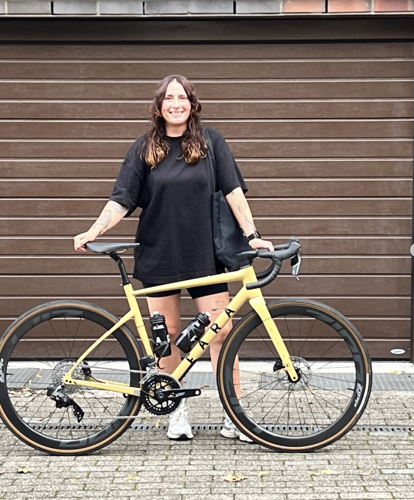 Eine lachende Frau mit dunklem, lockigem Haar, trägt schwarze Sportkleidung und steht neben einem gelben Fahrrad vor einer braunen, vertikalen Holzlamellenwand.