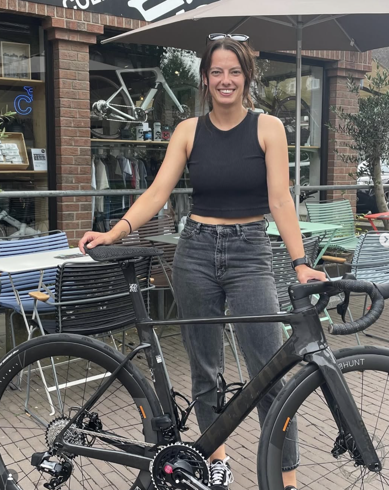 Eine lachende Frau mit braunen Haaren, Sonnenbrille auf dem Kopf, trägt einen schwarzen Tanktop und Jeans, steht neben einem schwarzen Rennrad vor einem Café mit mehreren Stühlen im Außenbereich.