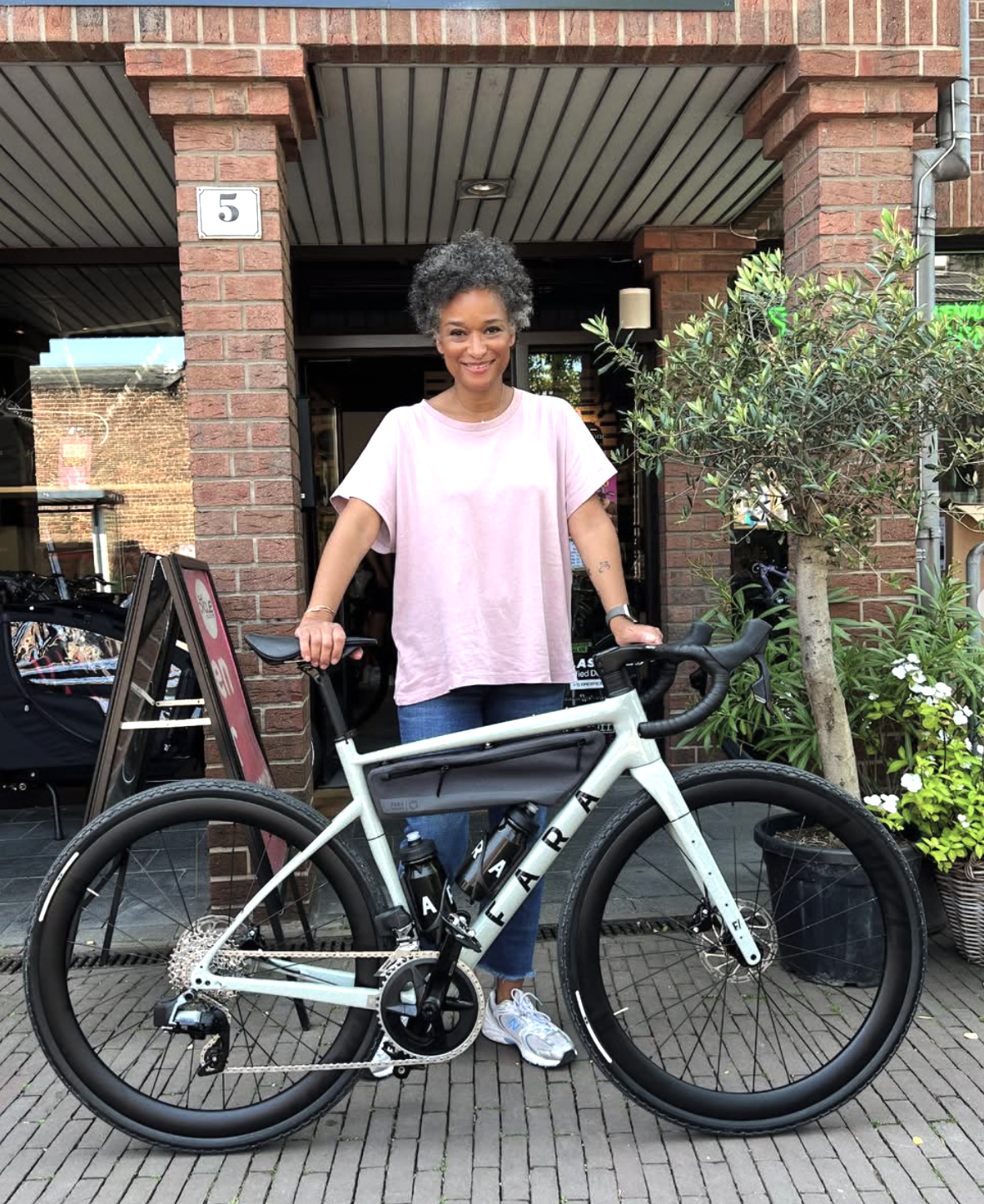 Eine Frau steht mit einem weißen Fahrrad vor einem roten Backsteingebäude, umgeben von Pflanzen und einem Baum.