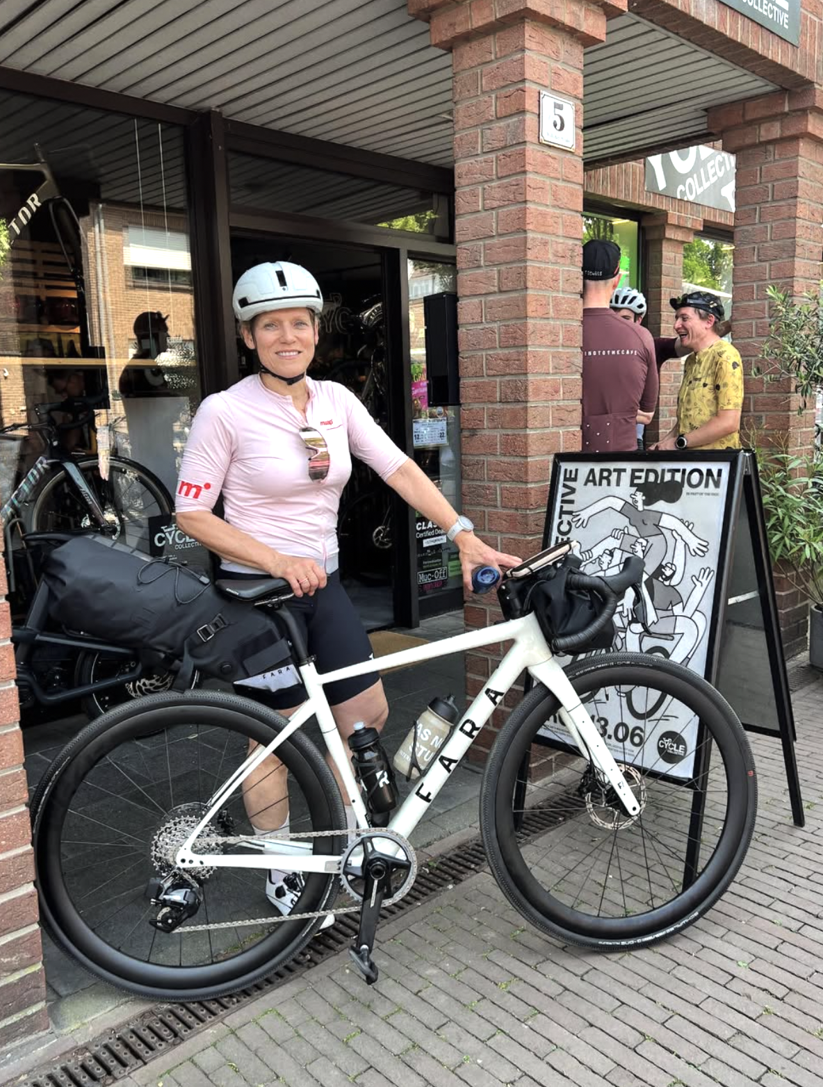 Eine Frau mit Fahrrad und Helm posiert vor einem Fahrradgeschäft. Im Hintergrund stehen mehrere Menschen, einige mit Helmen, und eine Litfaßsäule mit einem Kunstplakat.