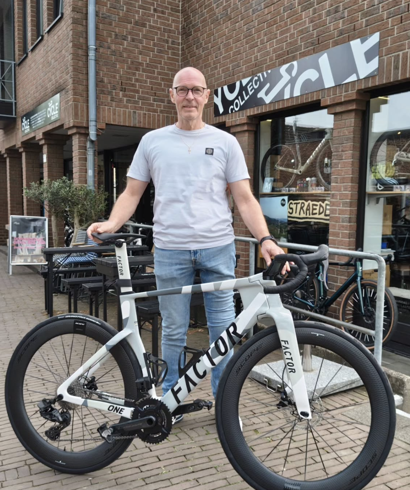 Ein älterer Mann mit Brillen steht vor einem Fahrradgeschäft und hält ein weißes Fahrrad der Marke Factor. Das Geschäft hat eine rote Backsteinfassade mit Schaufenstern, in denen Fahrräder und Fahrradzubehör zu sehen sind.