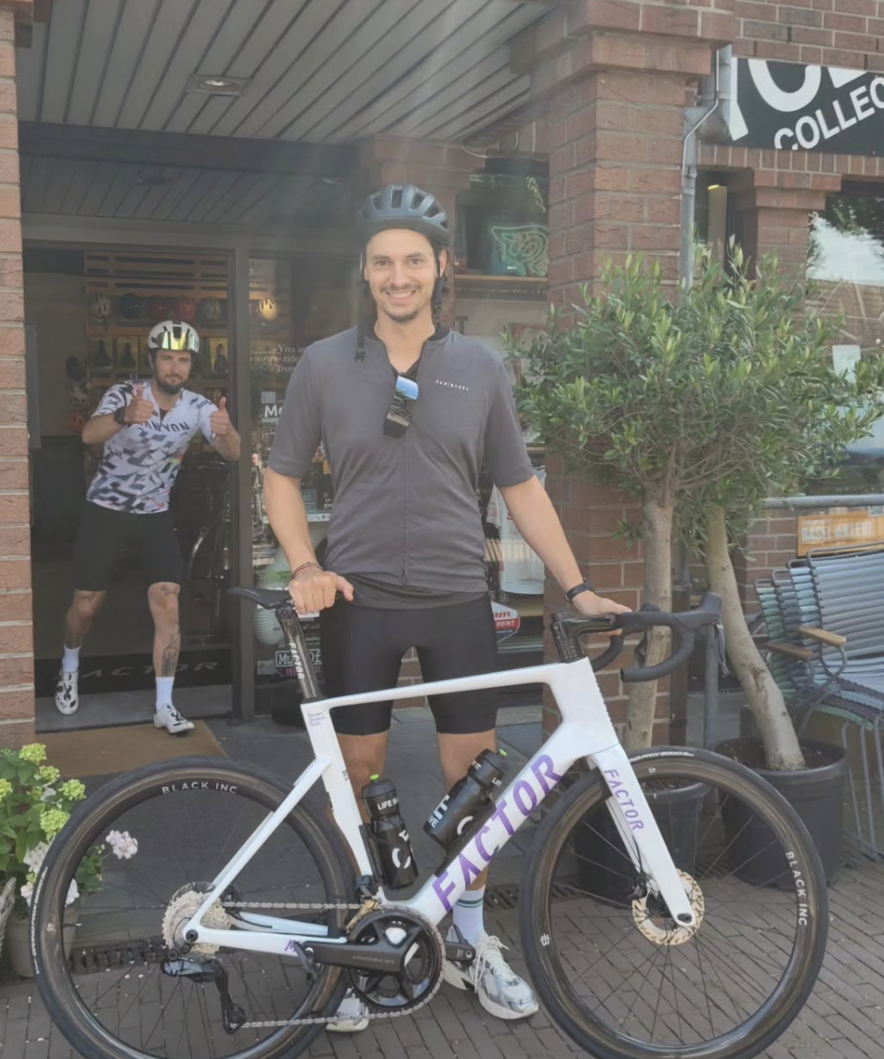 Zwei Männer, einer mit Fahrrad, beide tragen Fahrradhelme, vor einem Laden, einer neben einem weißen Fahrrad, der andere macht Daumen hoch im Hintergrund.