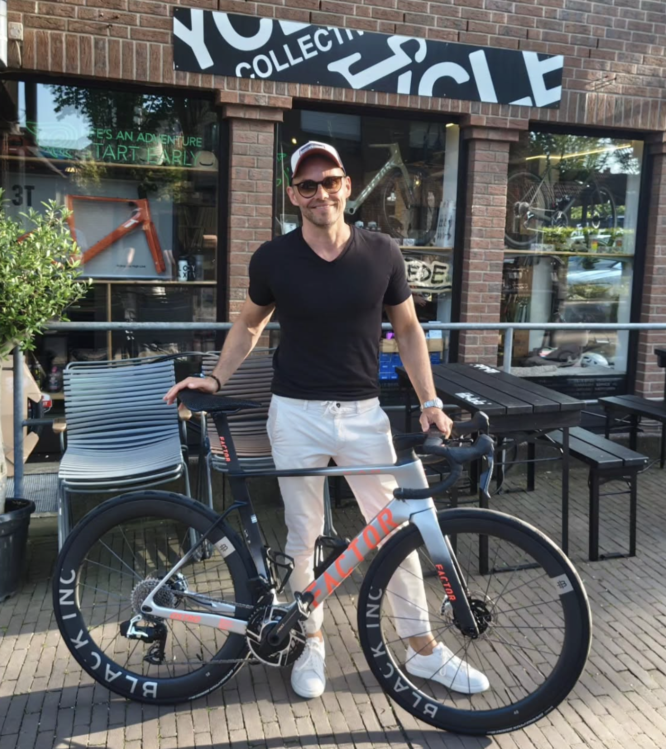 Mann steht mit Fahrrad vor einer Fahrradgeschäftsstrecke, trägt schwarze T-Shirt, helle Hosen, Sonnenbrille und eine Mütze, im Hintergrund eine Fensterfront mit Fahrradaccessoires.