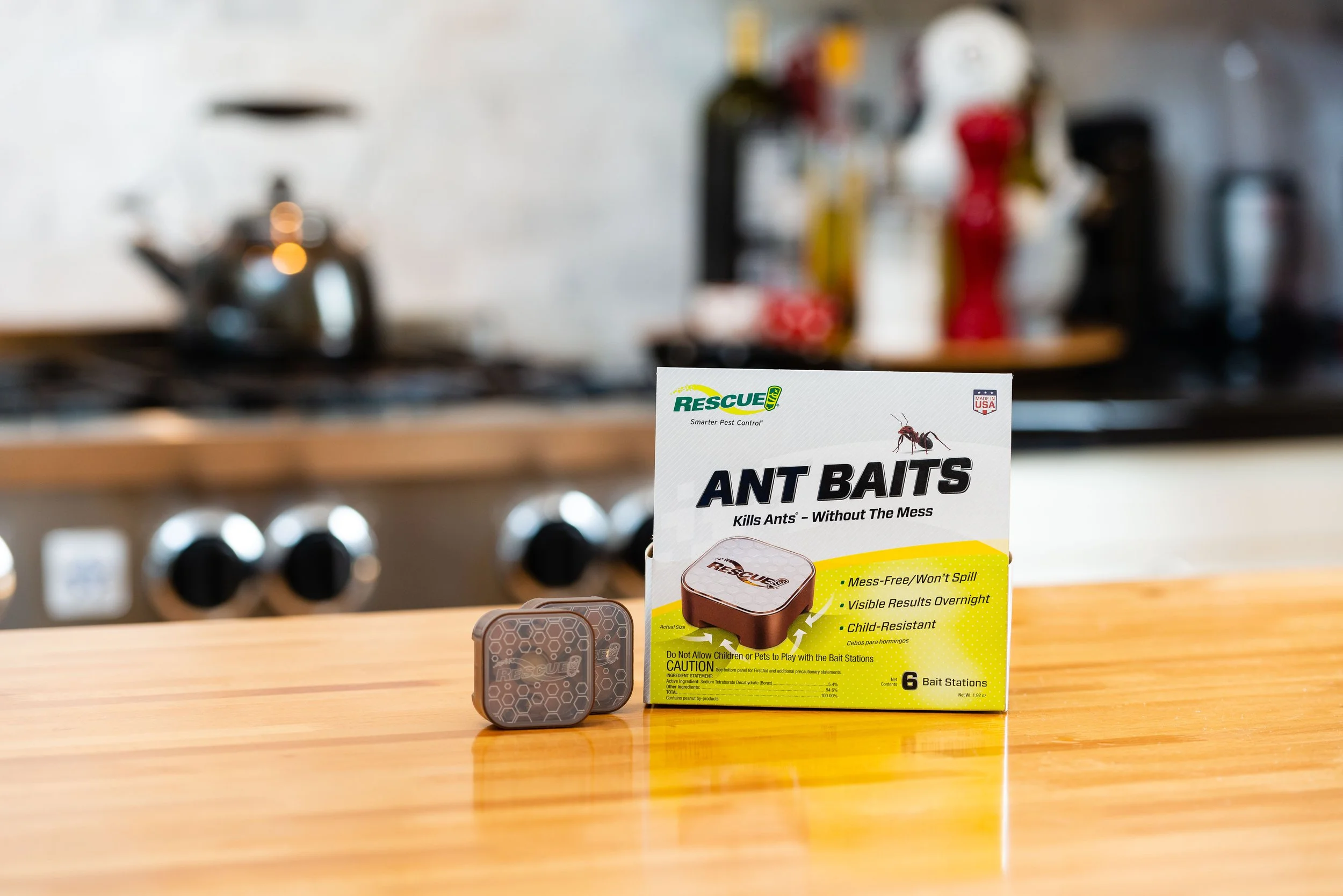 Ant bait product package on a wooden kitchen counter with a blurred stove and kitchen items in the background.