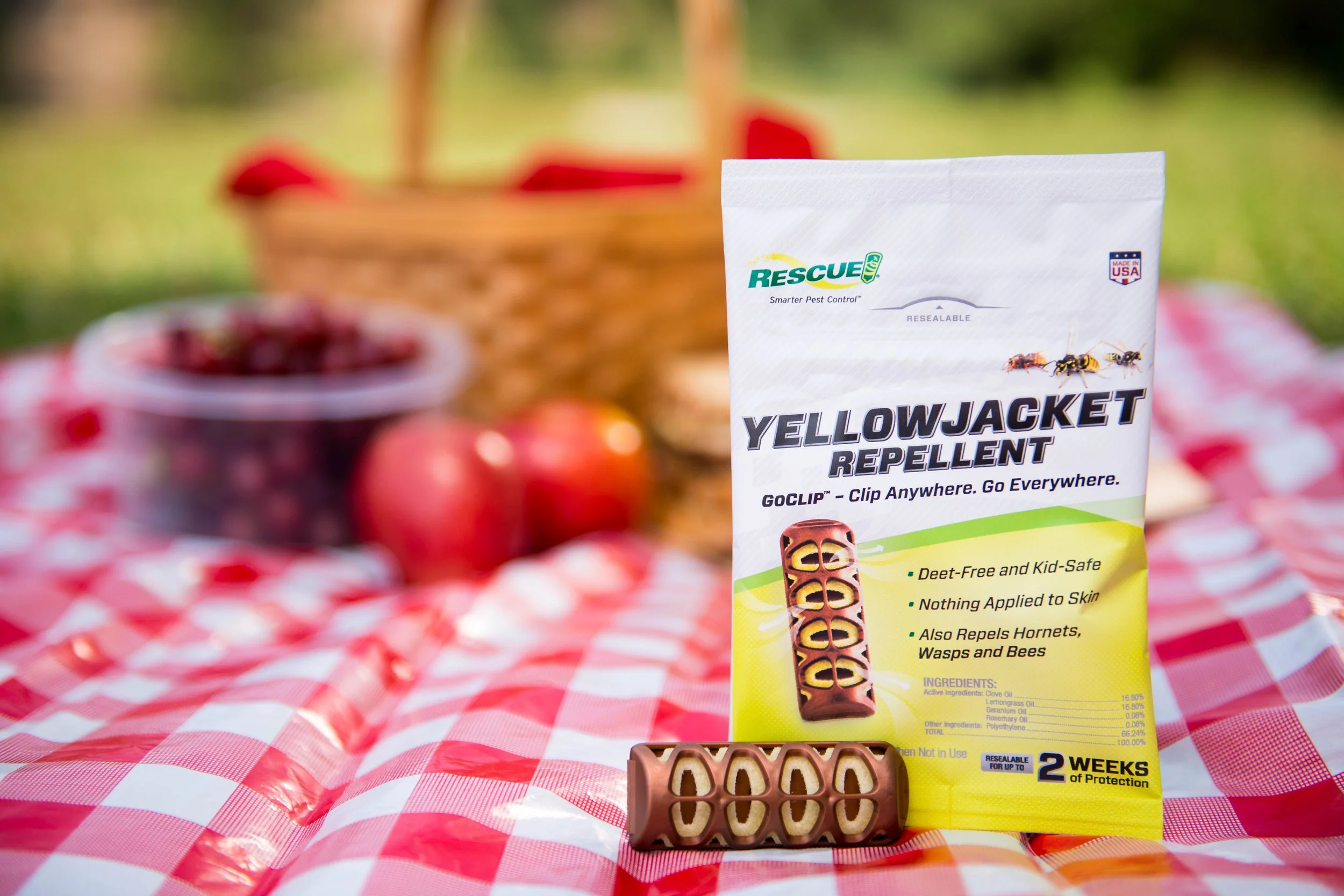 A package of Rescue Yellowjacket Repellent on a picnic table with a piece of chocolate-shaped pest-repellent candy in front, a bowl of cherries, and apples in the background. The scene is outdoors with a picnic basket and green grass.