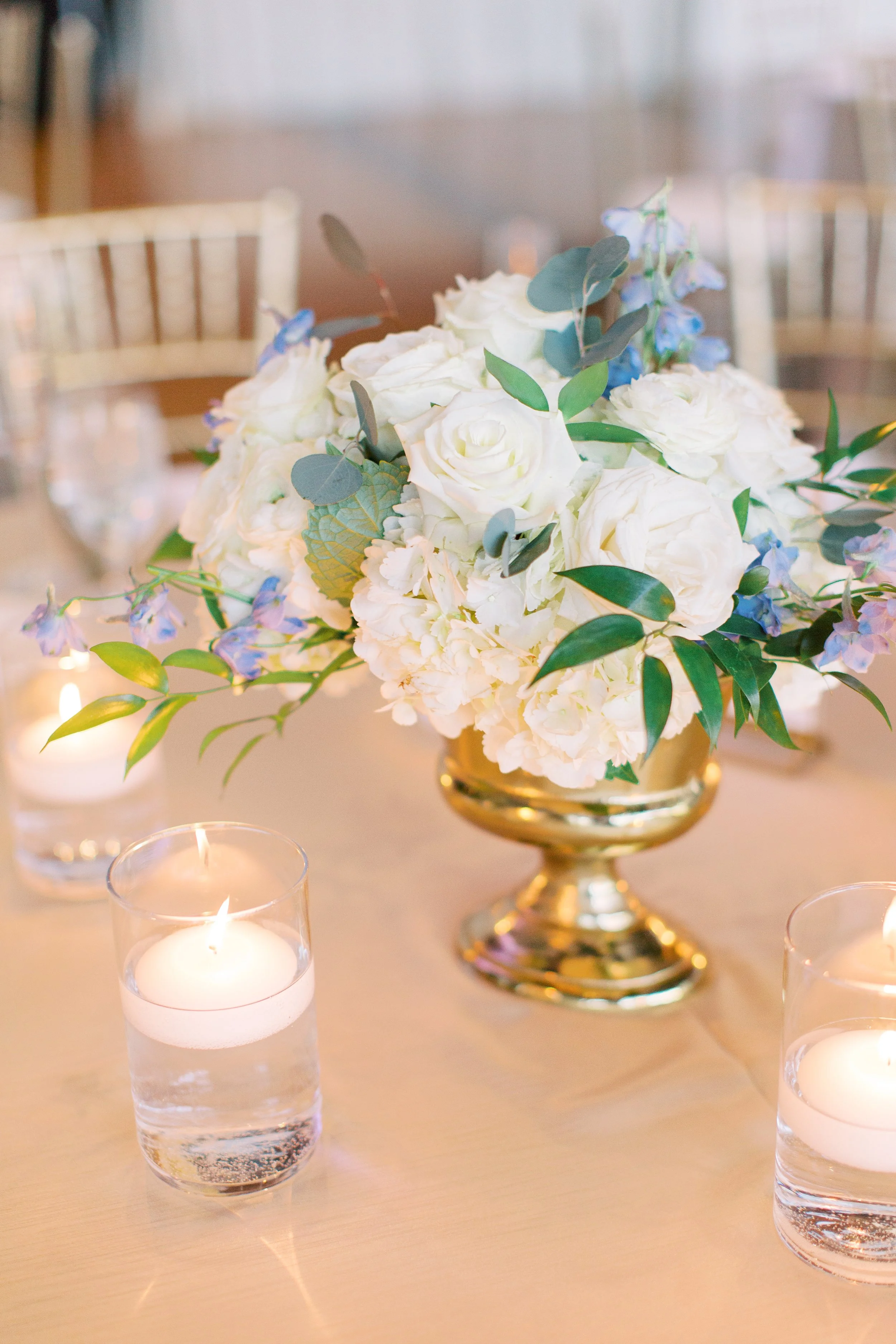 A floral centerpiece with white roses, hydrangeas, and eucalyptus leaves in a golden vase, surrounded by lit candles on a table.