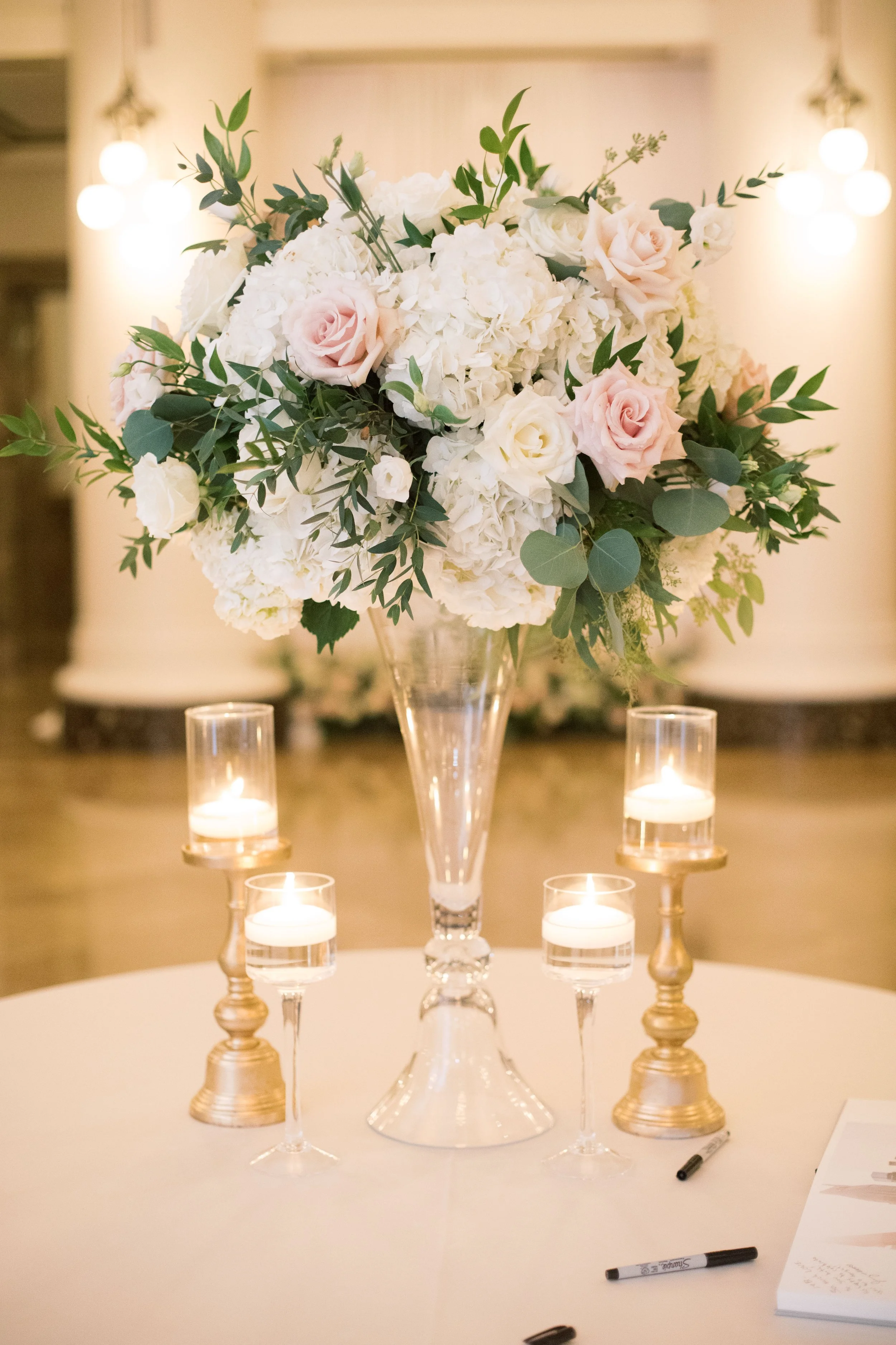 A floral centerpiece with white hydrangeas, light pink and white roses, and greenery in a tall glass vase, surrounded by four lit candles in glass holders on a round table.