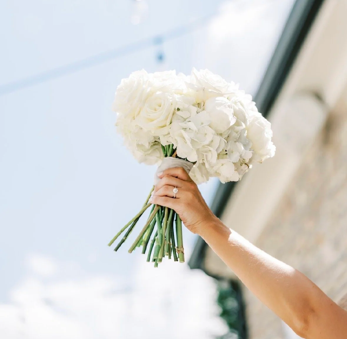 A person holding a bouquet of white flowers, including roses and hydrangeas, with an engagement ring on their finger, against a bright sky background.