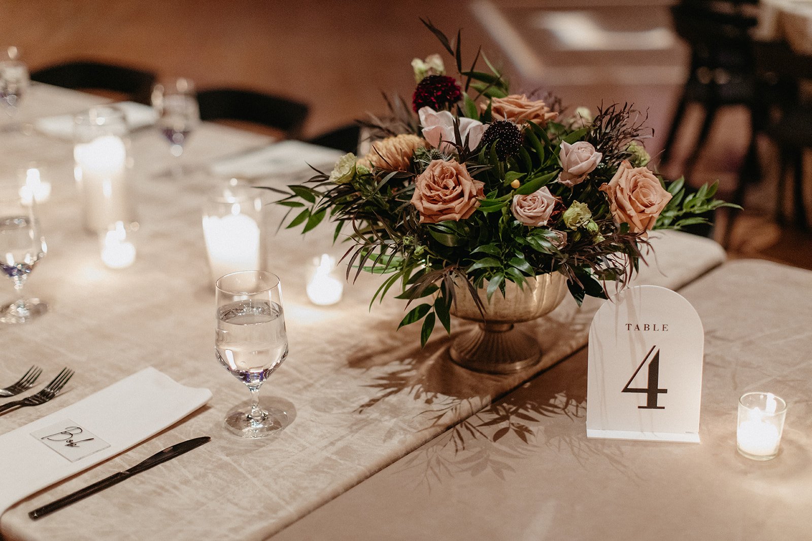 A wedding reception table decorated with a large floral centerpiece, lit candles, a water glass, and place settings with a napkin and utensils, with a table number sign reading 'Table 4'.