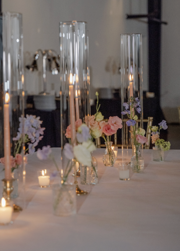 Elegant table setting with tall glass candle holders and small floral arrangements in vases, lit by candles, at an indoor event.