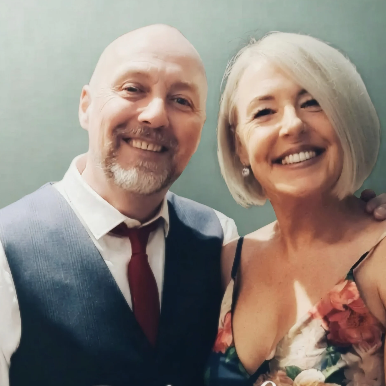 A middle-aged man with a bald head, beard, wearing a dress shirt, vest, and tie, smiling next to a woman with short, white hair, wearing a floral dress, both smiling and close together.