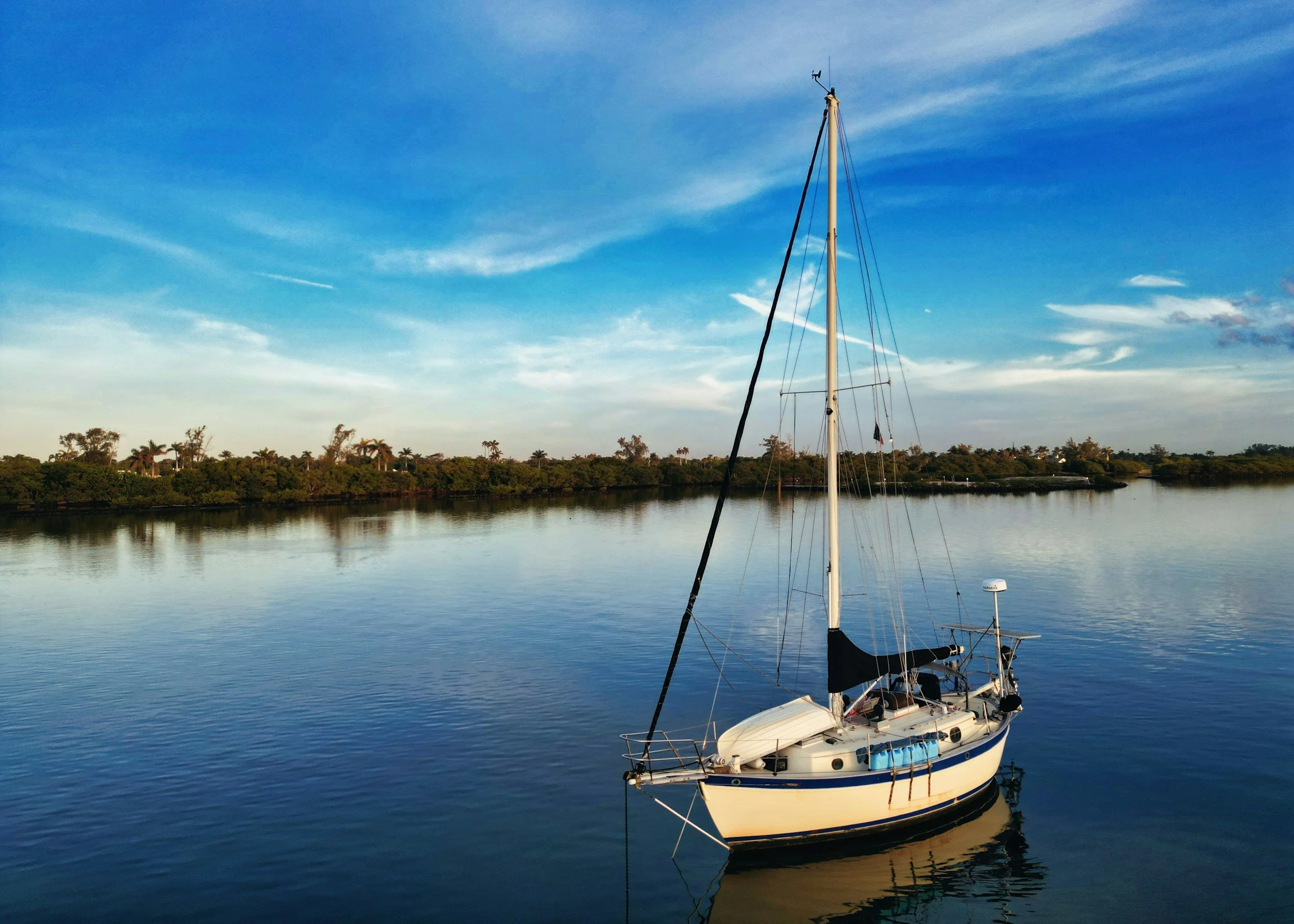 A white sailboat with a tall mast is anchored on calm, reflective water near a shoreline with trees. The sky above is blue with wispy clouds, creating a peaceful and serene scene.