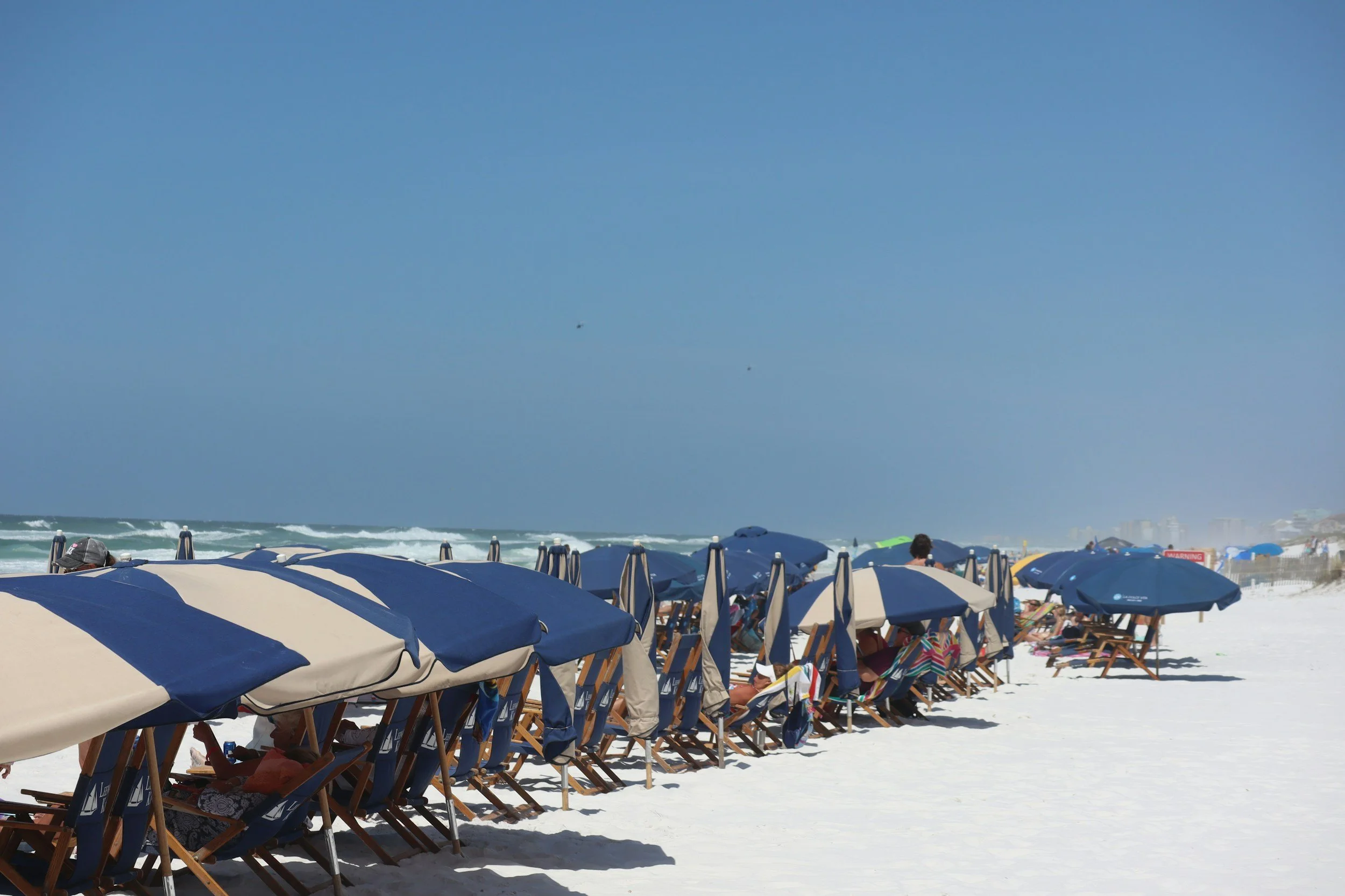 Rows of blue and beige beach umbrellas and chairs line a sandy beach, with people relaxing underneath. The ocean waves roll in the background under a clear blue sky.