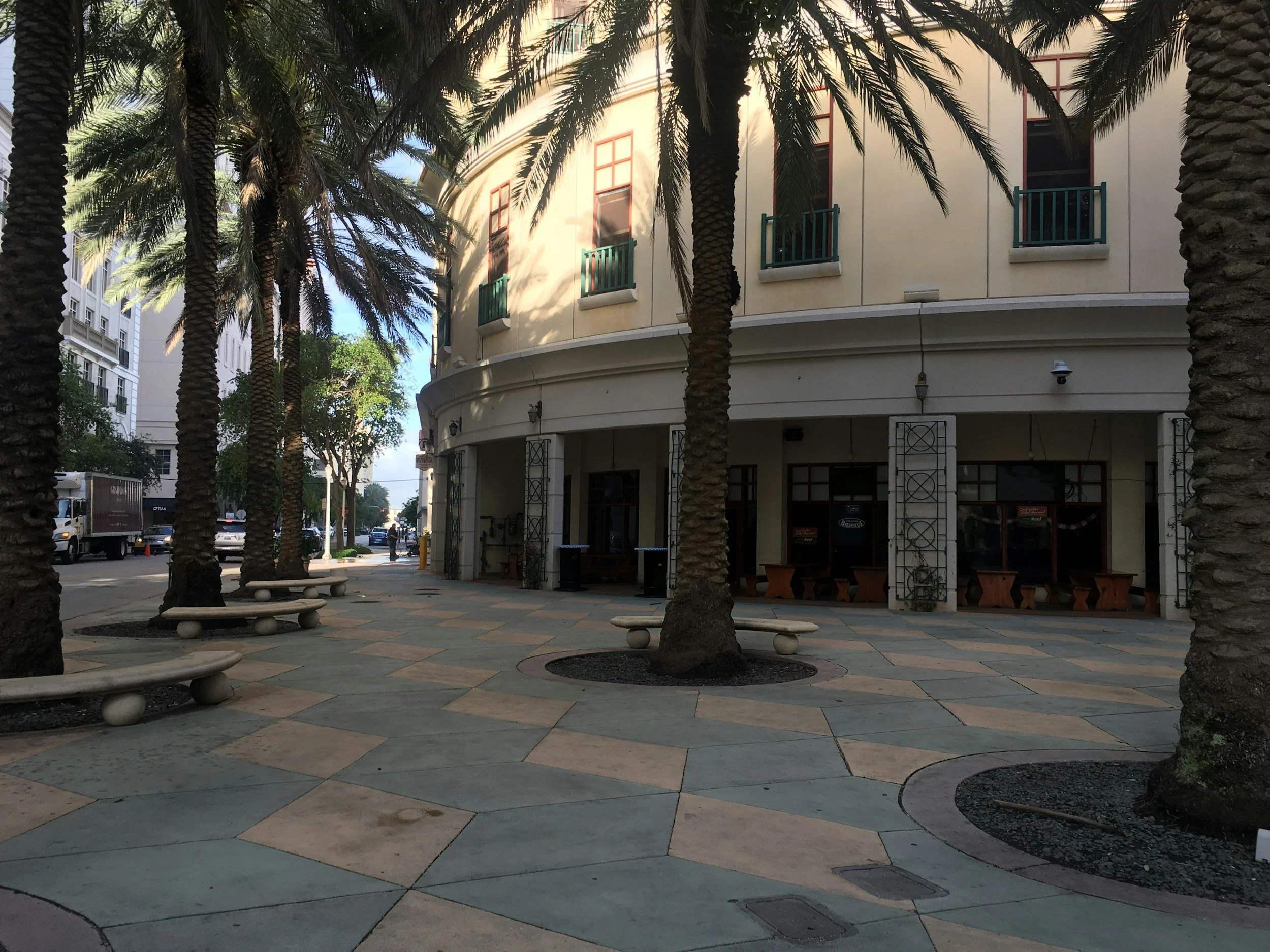 A sunlit sidewalk lined with palm trees curves around a light-colored building with green window railings. Benches sit beneath the trees, and shopfronts face the street.