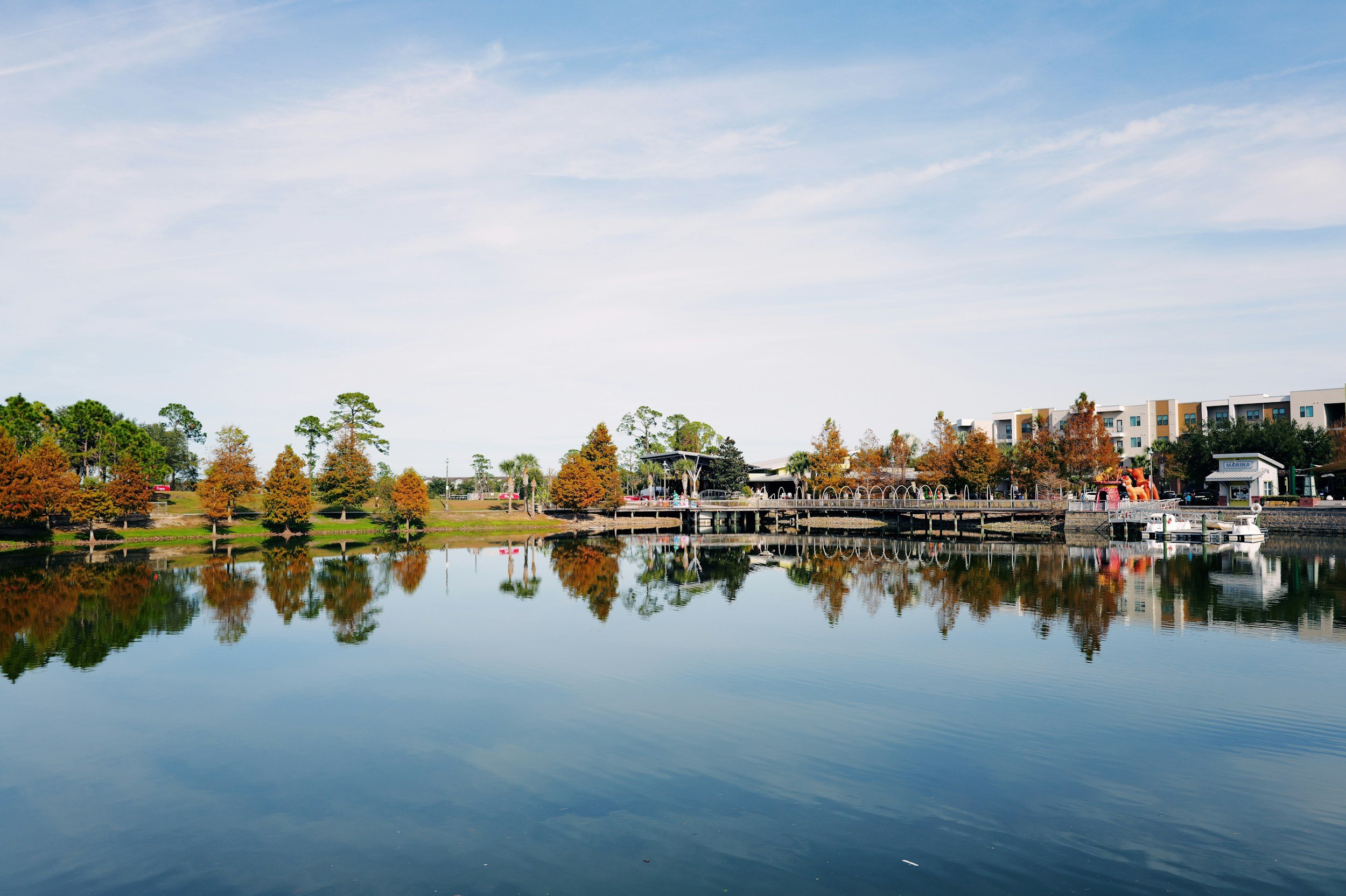 A calm lake reflects autumn trees, a wooden dock, and nearby buildings under a blue sky with scattered clouds.
