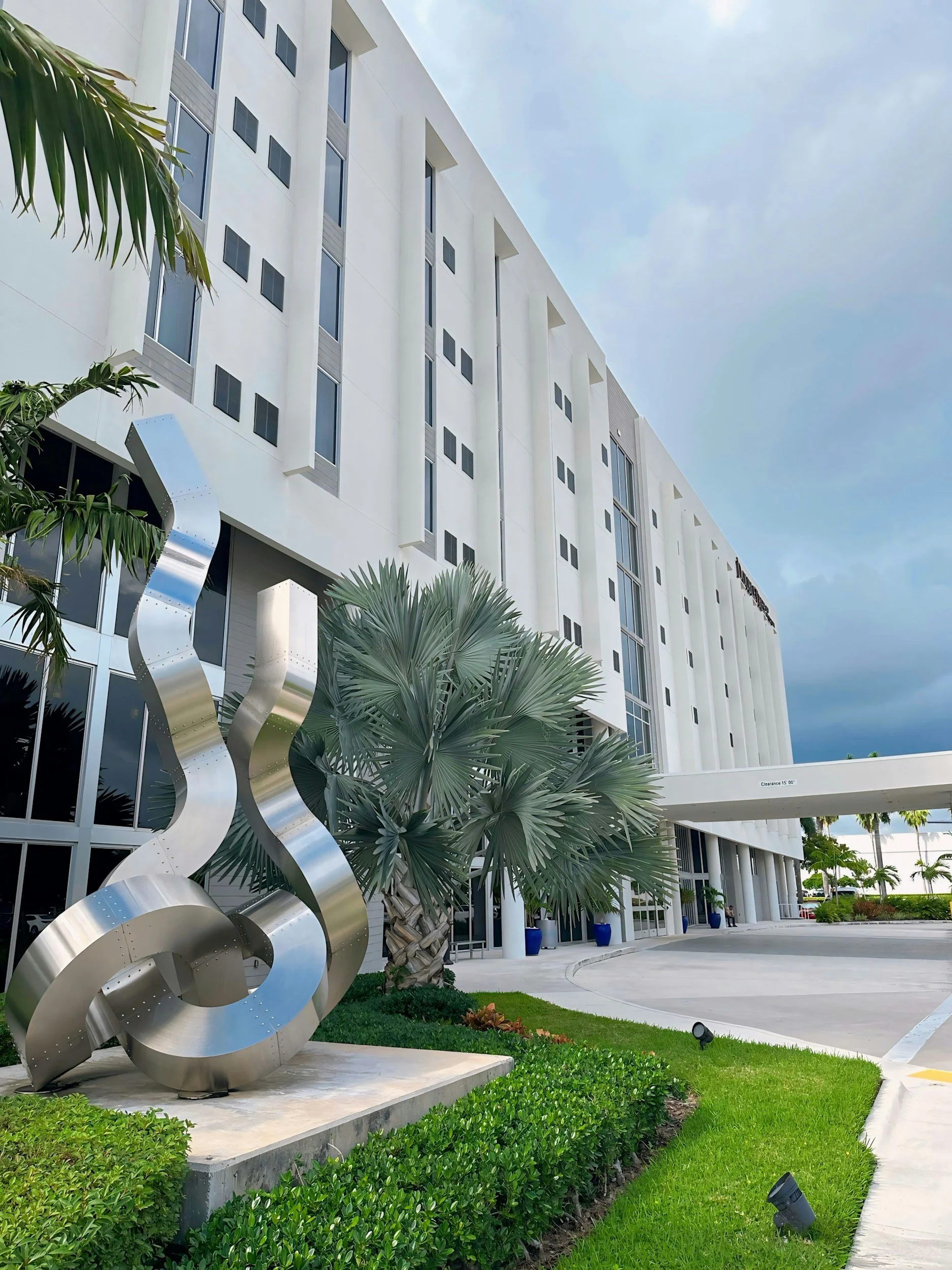 A modern white building with tall windows and a reflective silver abstract sculpture in front, surrounded by green plants and palm trees under a cloudy sky.