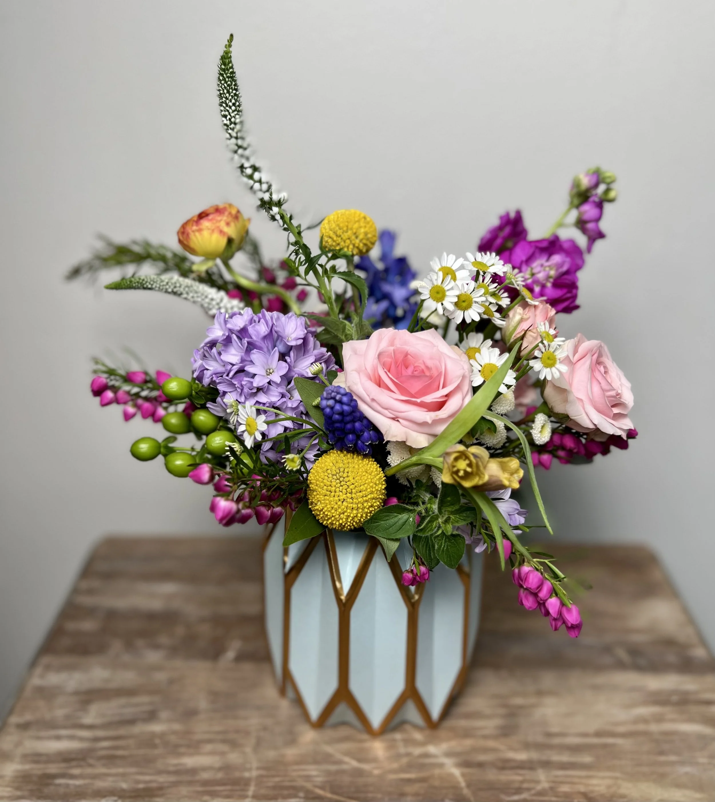 A colorful bouquet of various flowers in a white and gold geometric vase on a wooden table.