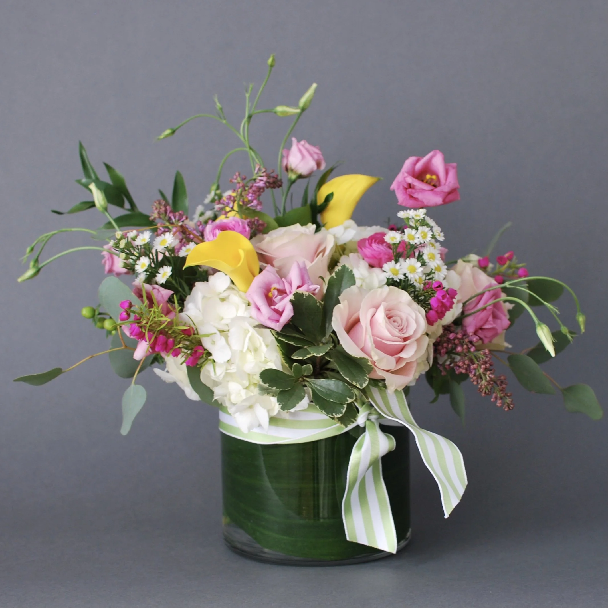 A floral arrangement in a green glass vase with pink roses, white hydrangeas, yellow calla lilies, pink lisianthus, daisies, and assorted greenery, accented with a striped ribbon.