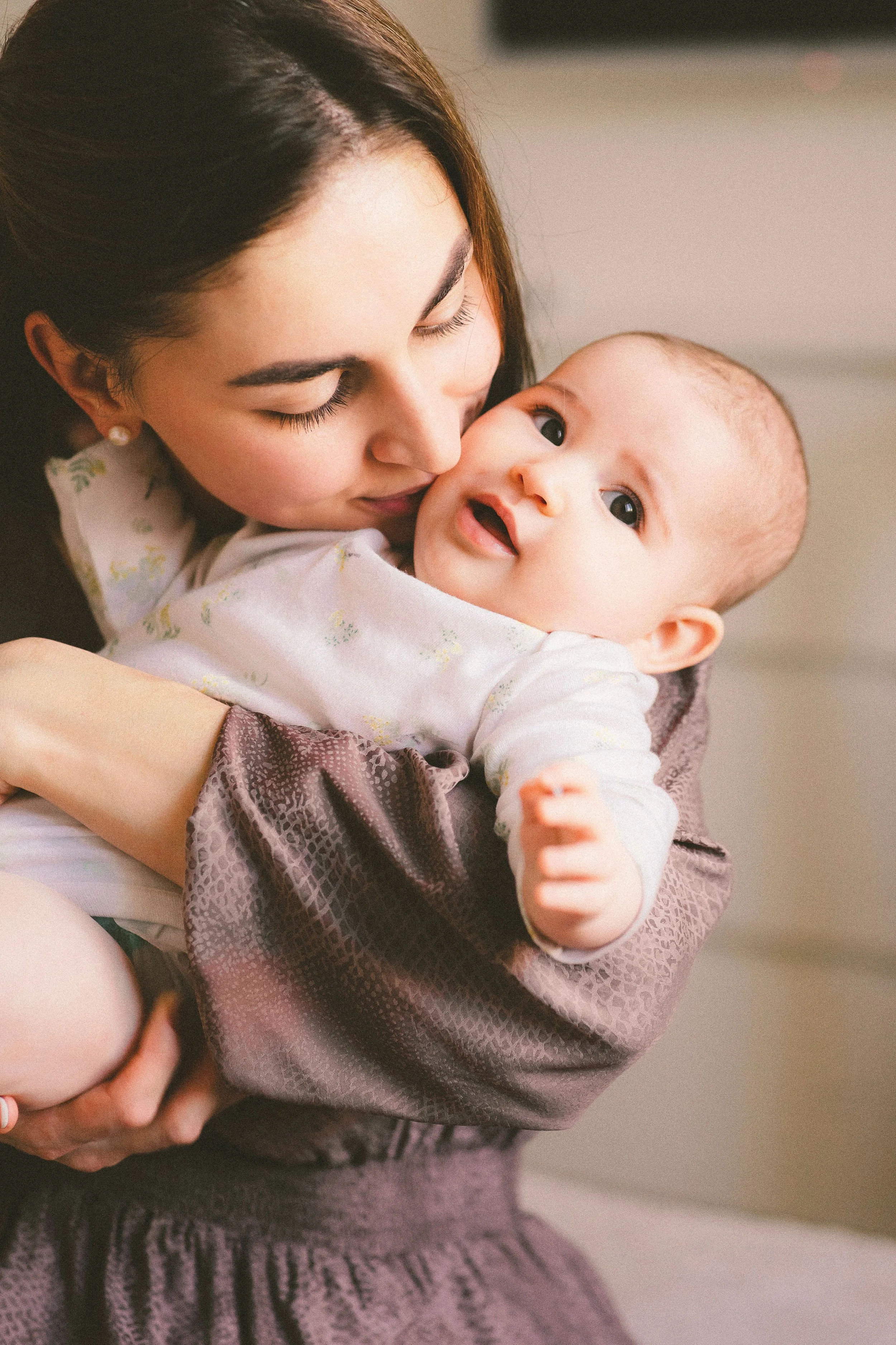 A mom with dark hair and earrings holds a baby with light hair, wide eyes, and a slightly open mouth, wrapped in a patterned blanket.