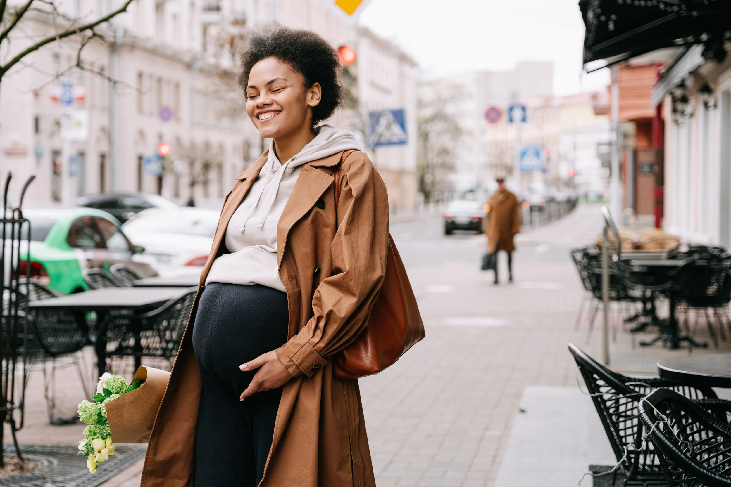 A smiling pregnant woman with curly hair, wearing a tan coat over a gray hoodie, stands on a city sidewalk holding a bouquet of green and white flowers in a brown paper bag.