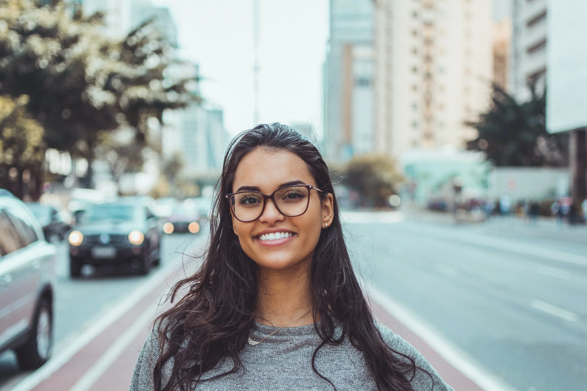 Smiling woman with long dark hair and glasses standing outdoors on a city street with tall buildings and trees in the background.
