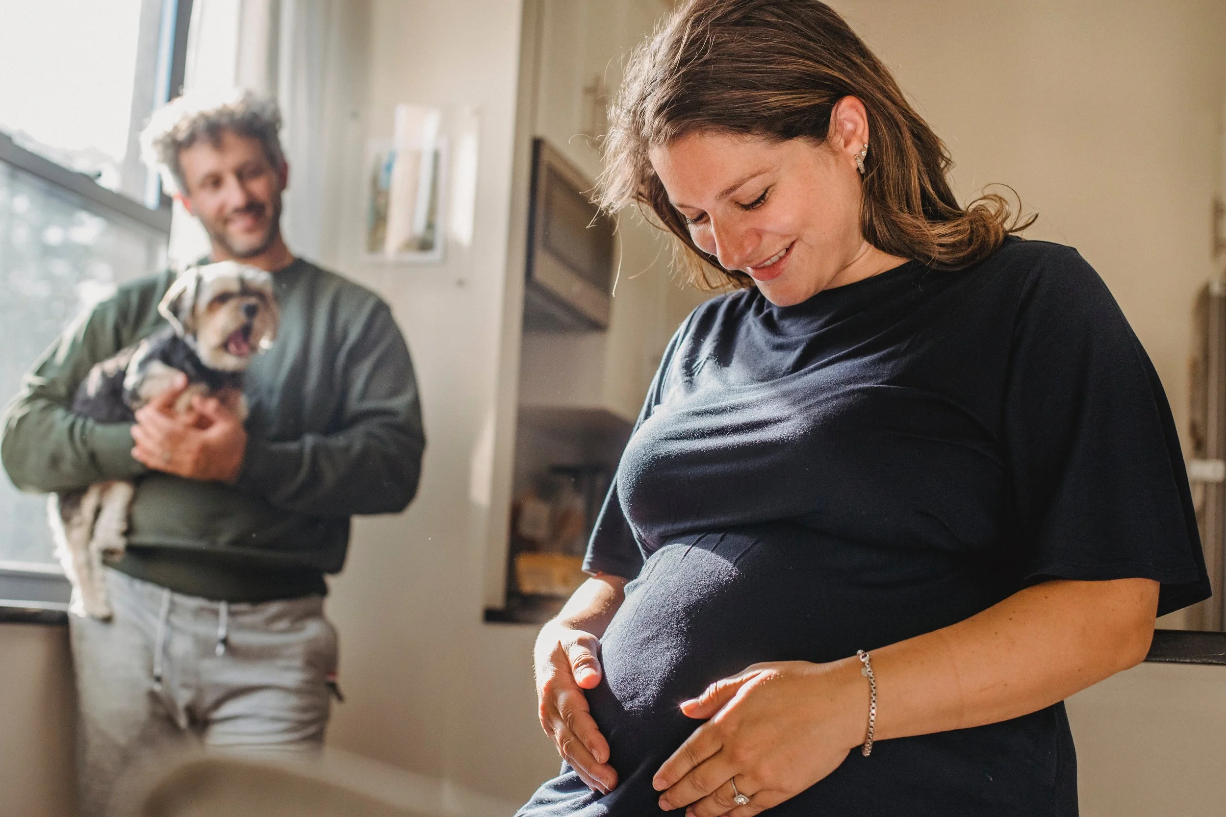 A mom is smiling and gently touching her pregnant belly, while a dad is in the background is holding a small puppy and smiling.