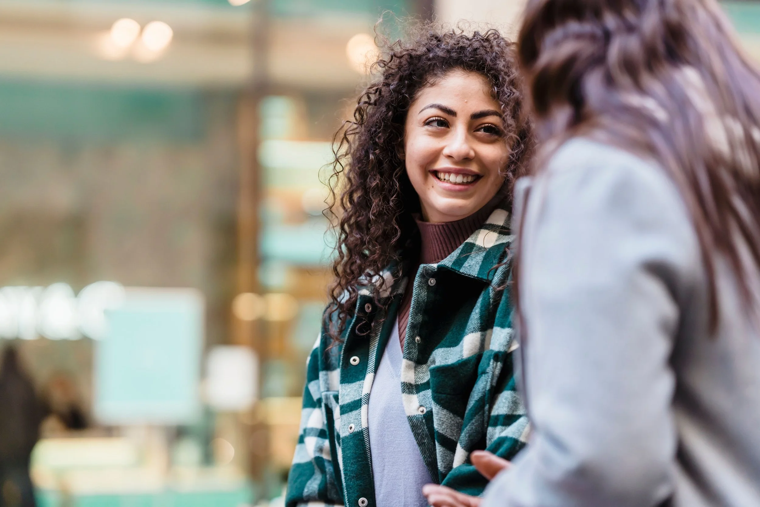 Two young women talking and smiling in an indoor setting, one with curly hair wearing a plaid shirt.