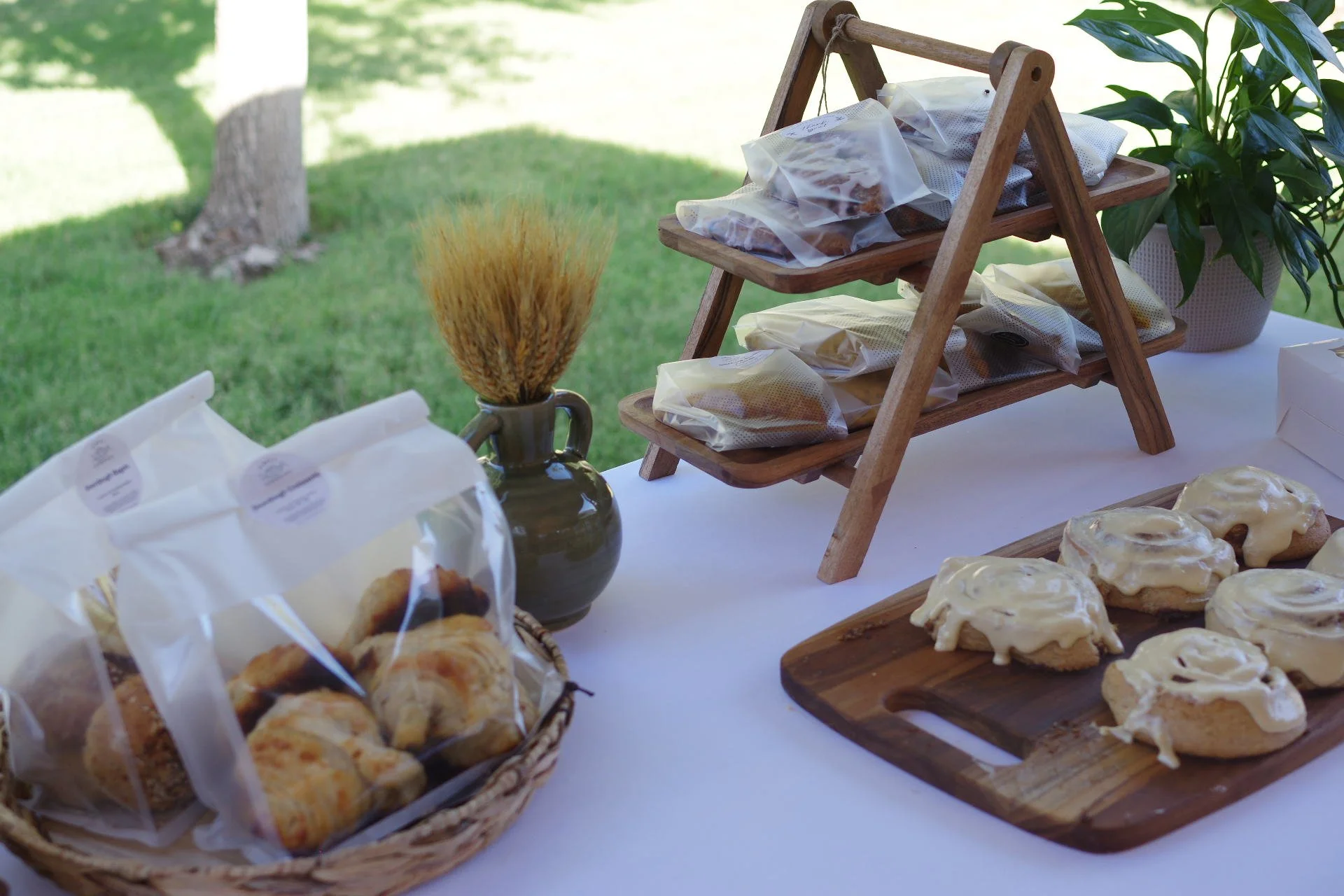Assortment of packaged sourdough pastries on display, including croissants, cookies, and cinnamon rolls, on a wooden stand and table, with a vase of wheat and a potted plant in the background.