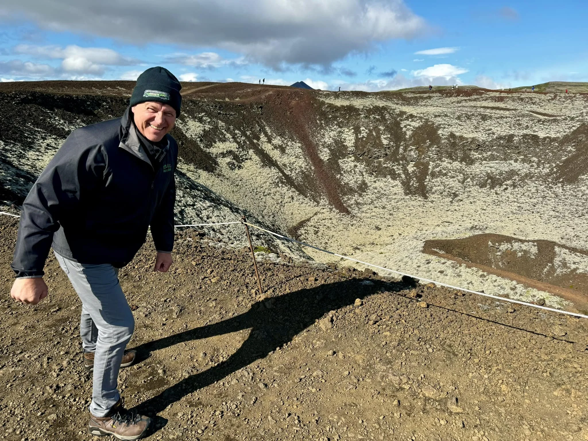 A smiling man wearing a black jacket, gray pants, hiking boots, and a navy beanie stands on a rocky, volcanic landscape near a crater with a white rope fence. The background features a large volcanic crater, partly cloudy sky, and distant people on the crater rim.
