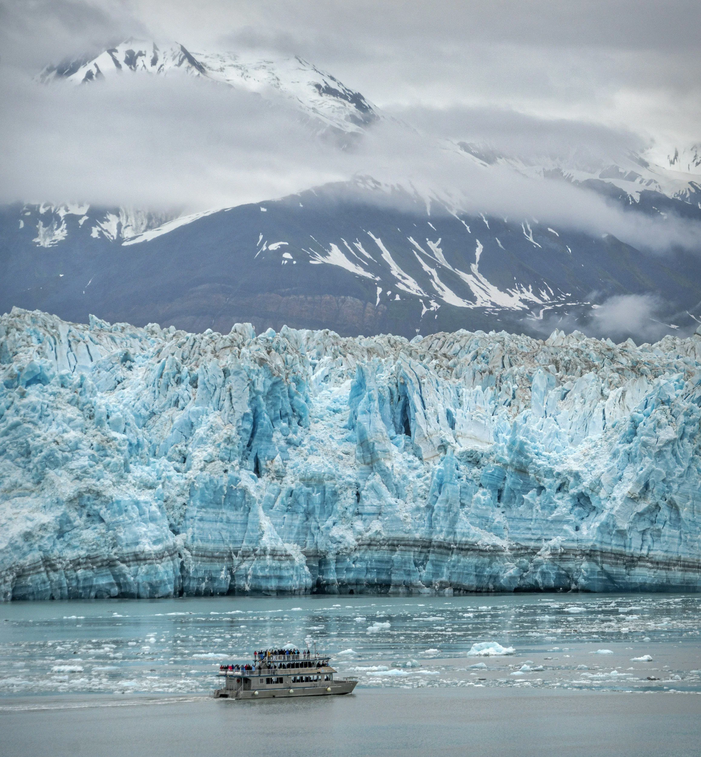 A boat with people on it navigates near floating icebergs and a glacier under snowy mountains shrouded in clouds.