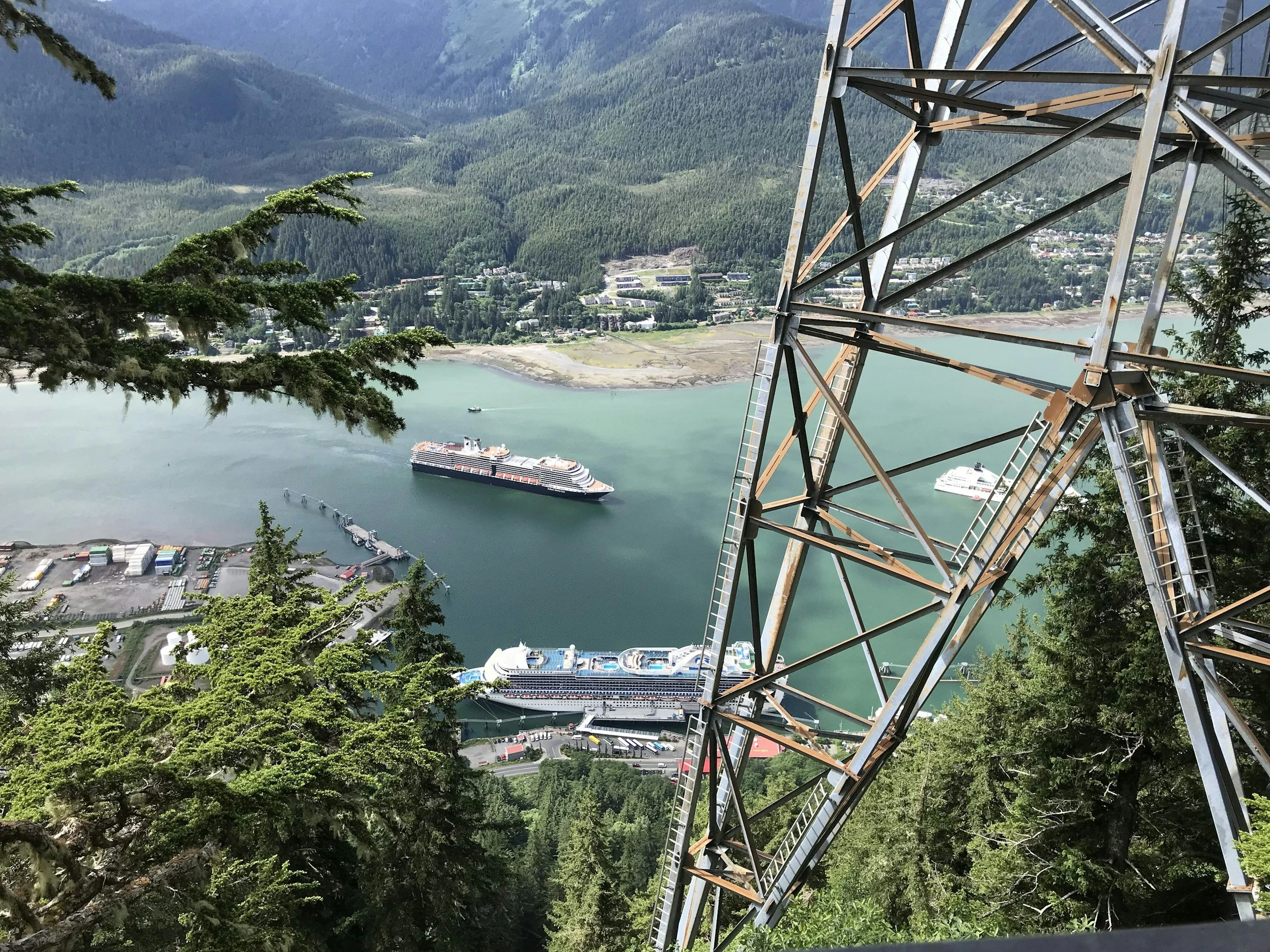 View from a high height of a large amusement park or cruise terminal with boats docked on the water, surrounded by lush green mountains and trees, with a section of a steel structure or ride in the foreground.