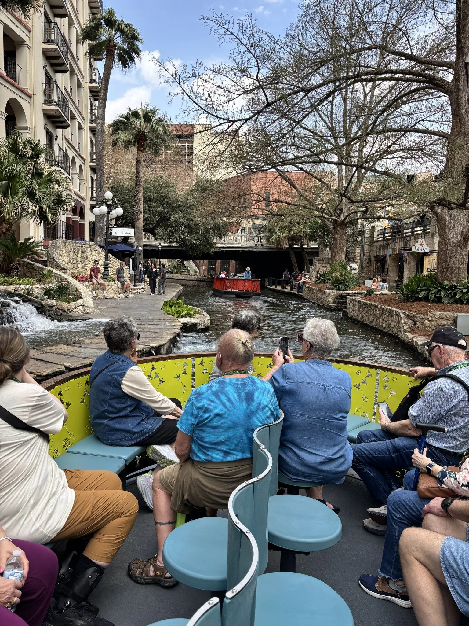 People seated on a boat tour along a river in an urban area with palm trees, a bridge, and buildings visible in the background.