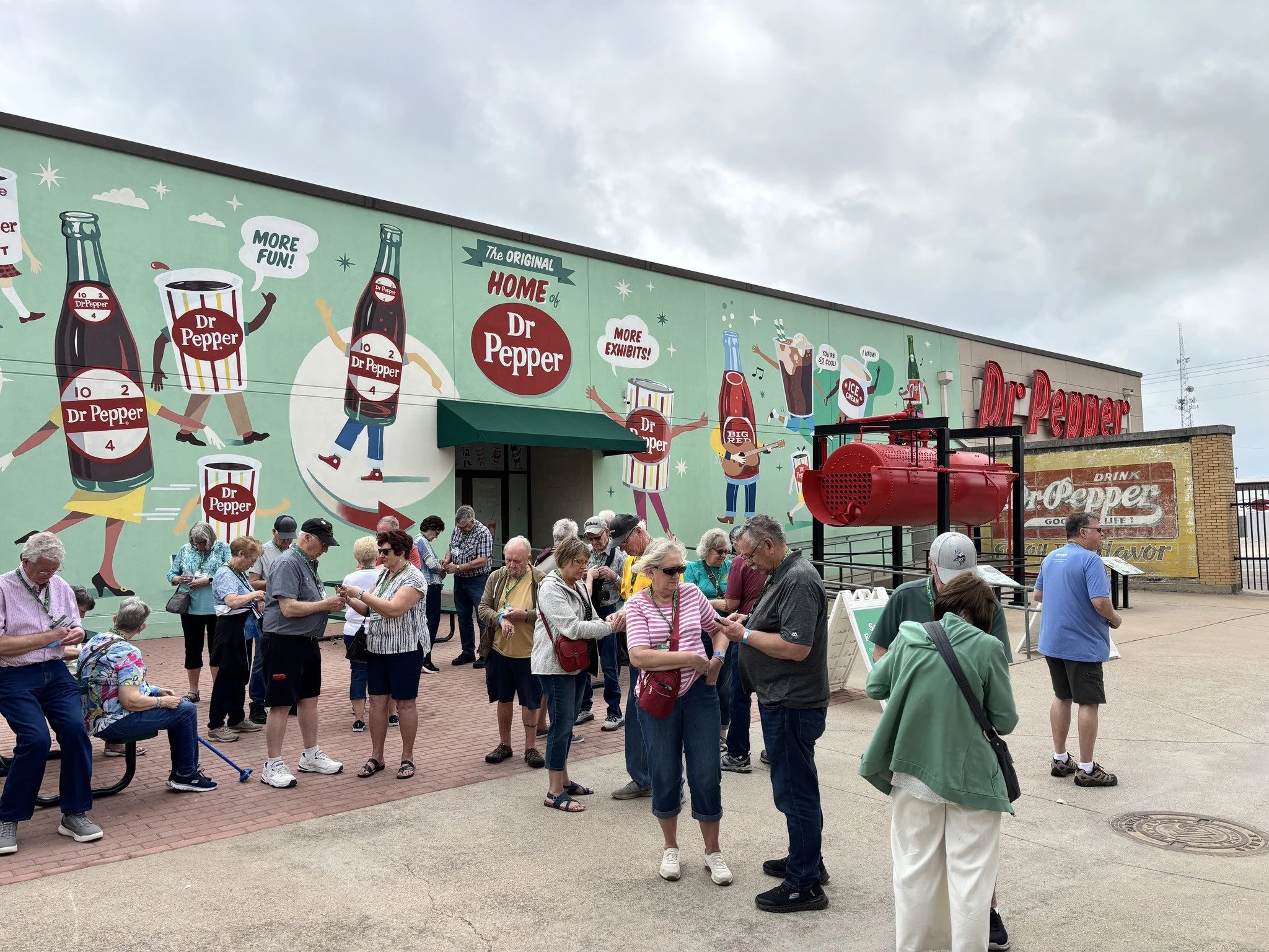 People waiting in line outside a Dr. Pepper store with a colorful outdoor mural featuring cartoon Dr. Pepper bottles and cans and the words "Home of Dr. Pepper."