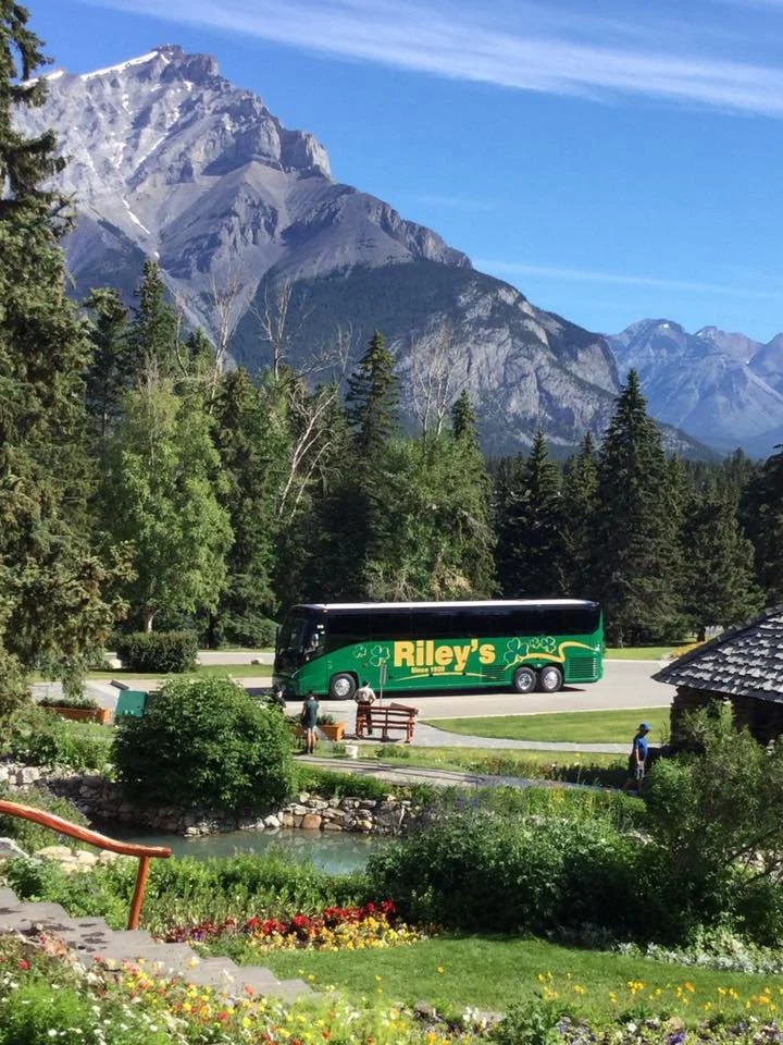 Tour bus with Riley's branding parked in a scenic mountain area with trees, a pond, and colorful flowers in the foreground and tall mountains in the background.