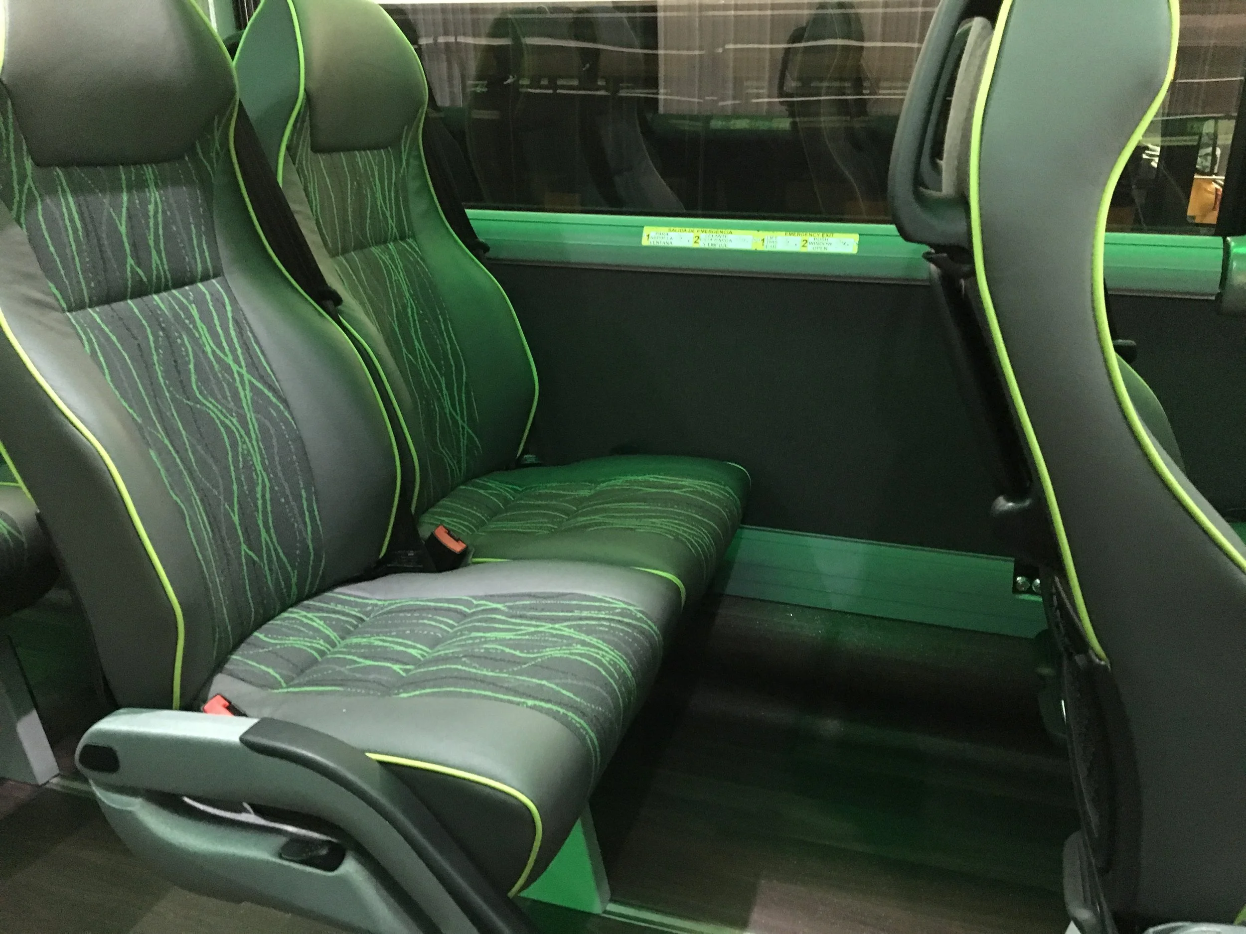 Inside a bus, green and black seats with green piping and patterned fabric, some seats are facing forward, and a window at the back.