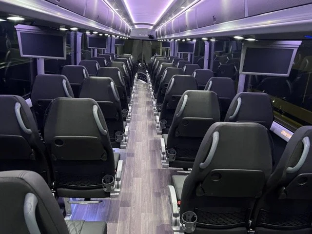 Interior of a modern bus or coach with rows of black seats, purple ambient lighting, and multiple screens on the walls.