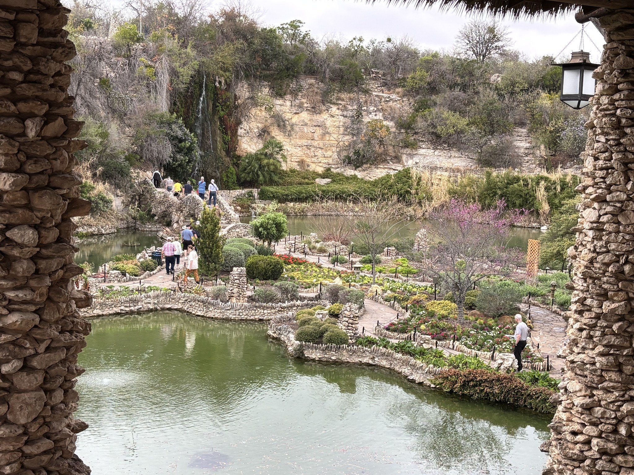 Scenic view of a pond surrounded by landscaped greenery and colorful flowers, with a rocky cliff in the background and several people walking along a path, viewed through a stone archway with a hanging lantern.