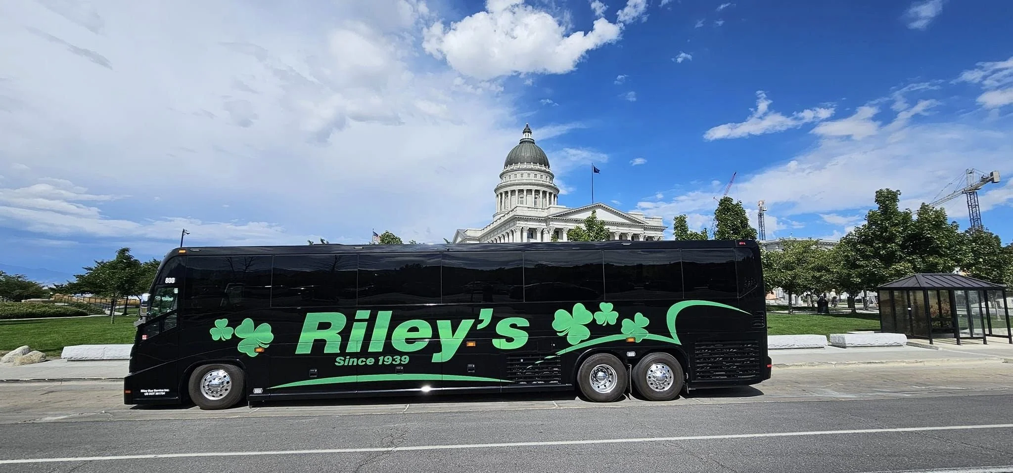 A black bus with green lettering and shamrock decorations parked on a street in front of a government building. The building is white with a dome, likely the Capitol, under a partly cloudy sky.