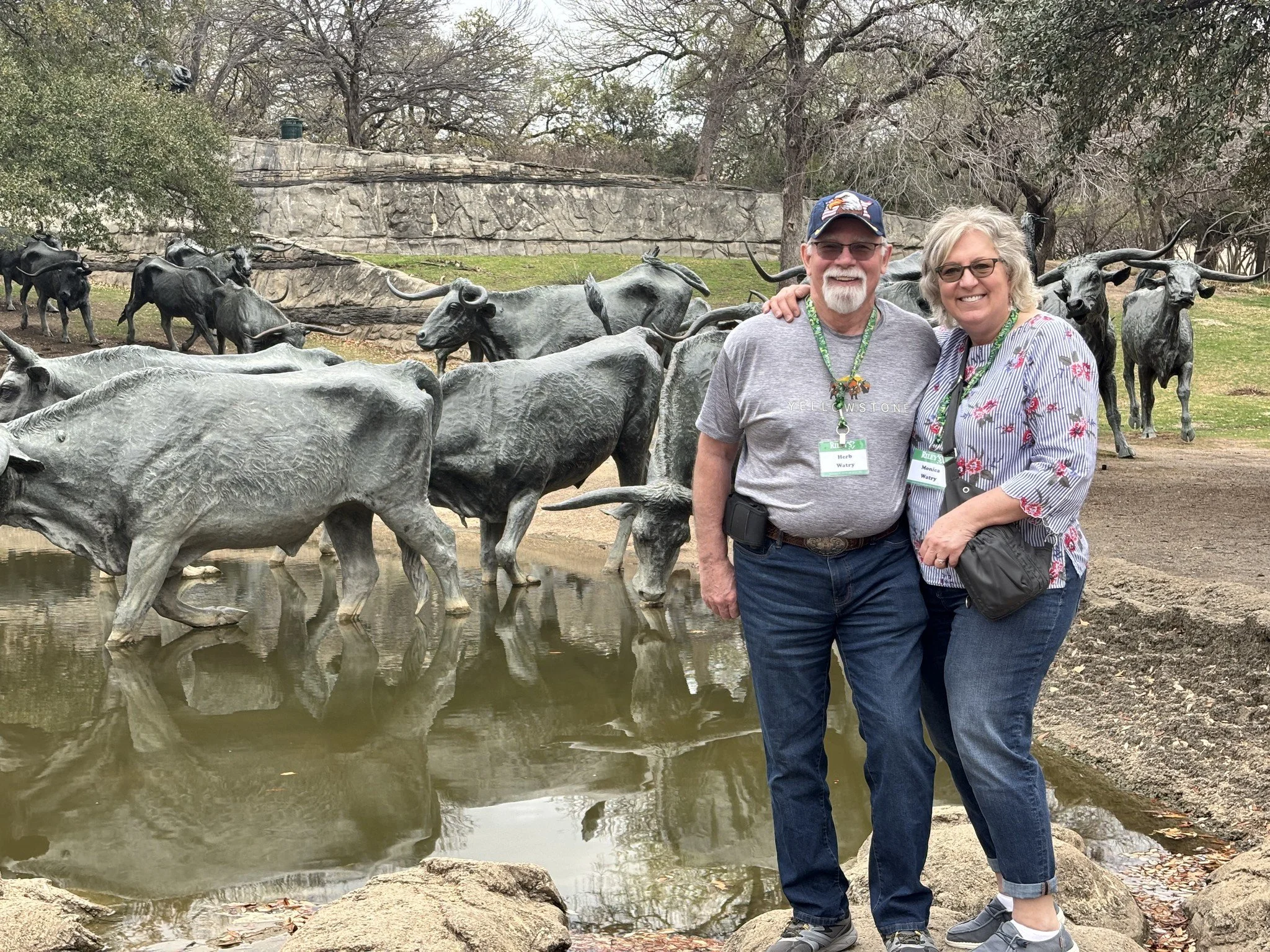 Two smiling people, a man and a woman, standing on rocks next to a pond with bronze sculptures of longhorn cattle, trees, and a stone wall in the background.