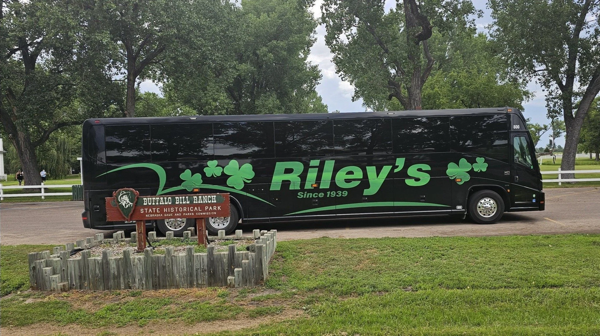 Black bus with green shamrock and lettering for Riley's, parked near a sign for Buffalo Bill Ranch State Historical Park, with trees and a white fence in the background.
