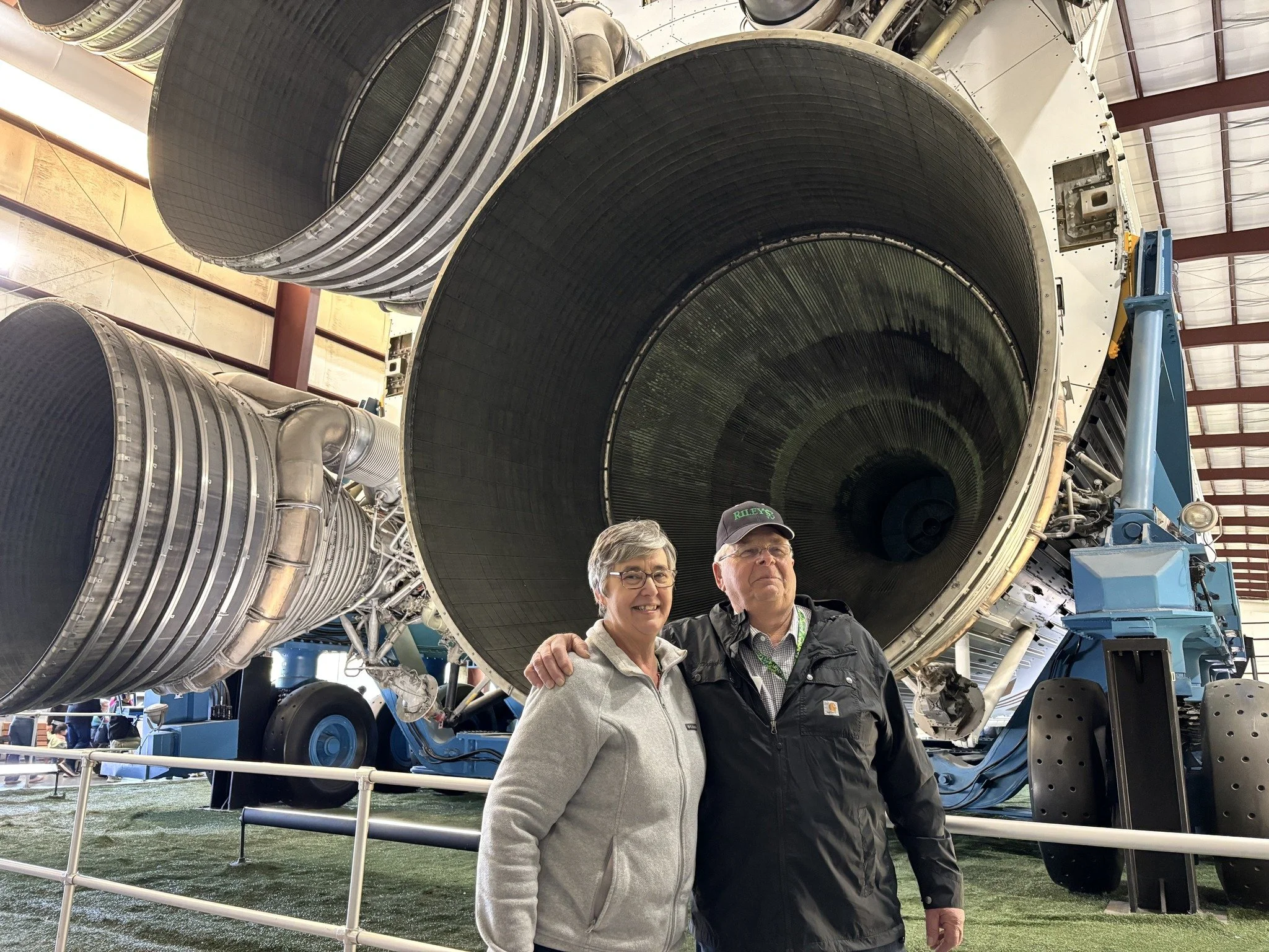 Two people, a woman and a man, standing in front of a large rocket engine at an aerospace exhibition. The engine has multiple stages with visible metal and tubing, and they are smiling.