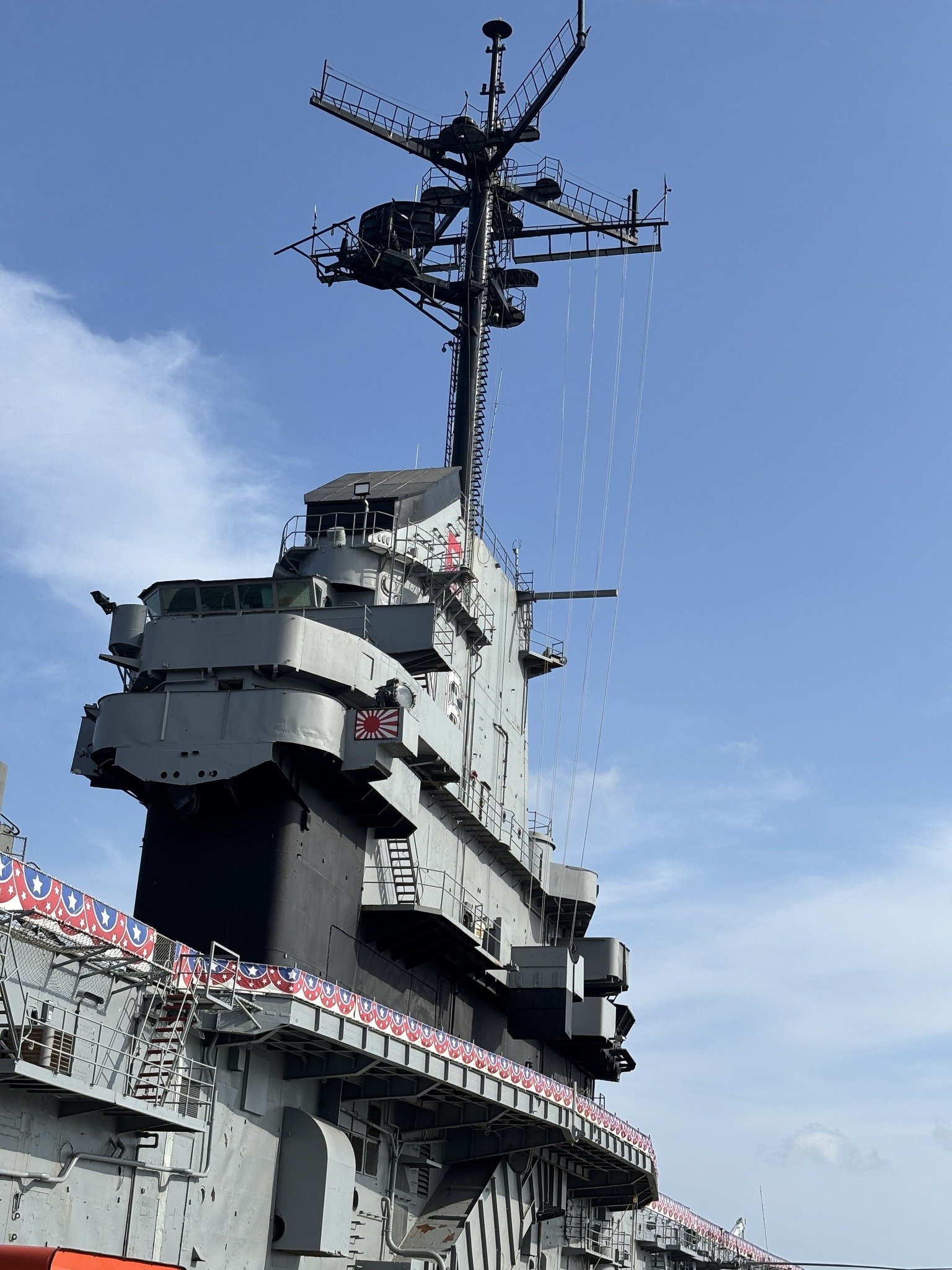 A close-up view of the superstructure and mast of a naval aircraft carrier against a partly cloudy blue sky, with patriotic bunting hanging along the ship's railings.