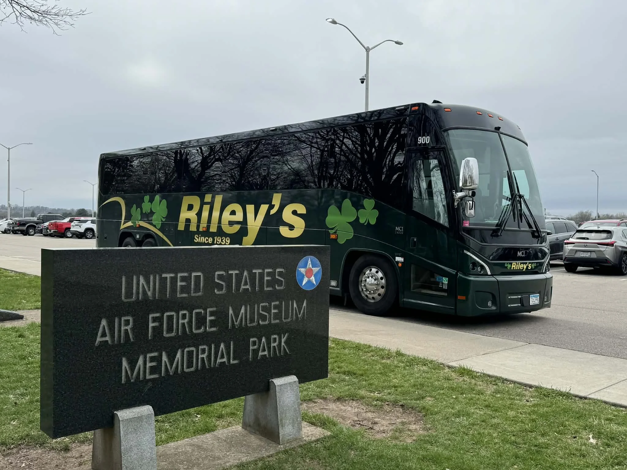 A black bus with green shamrock graphics and the label Rileys, parked near a sign for the United States Air Force Museum Memorial Park, with a parking lot of cars in the background and cloudy sky above.