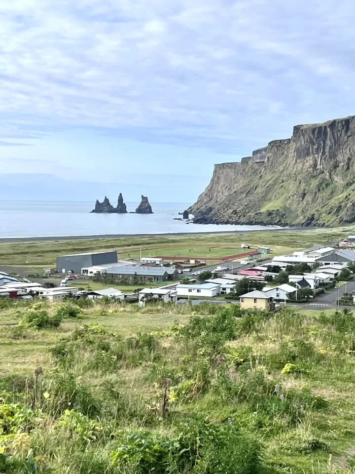 Scenic coastal village with white houses, green fields, a mountain with cliffs, and sea stacks in the ocean on a partly cloudy day.